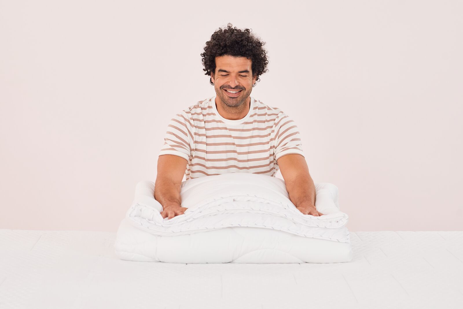 Persona seduta su un letto mentre piega un piumone bianco, con maglietta a righe beige e bianche.