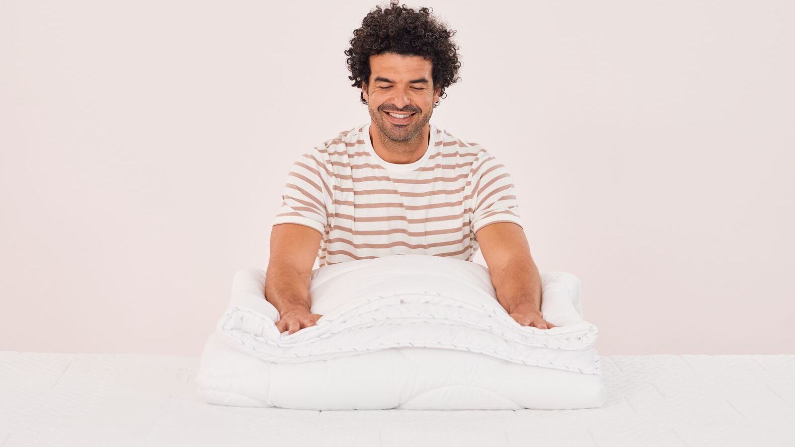 Persona seduta su un letto mentre piega un piumone bianco, con maglietta a righe beige e bianche.