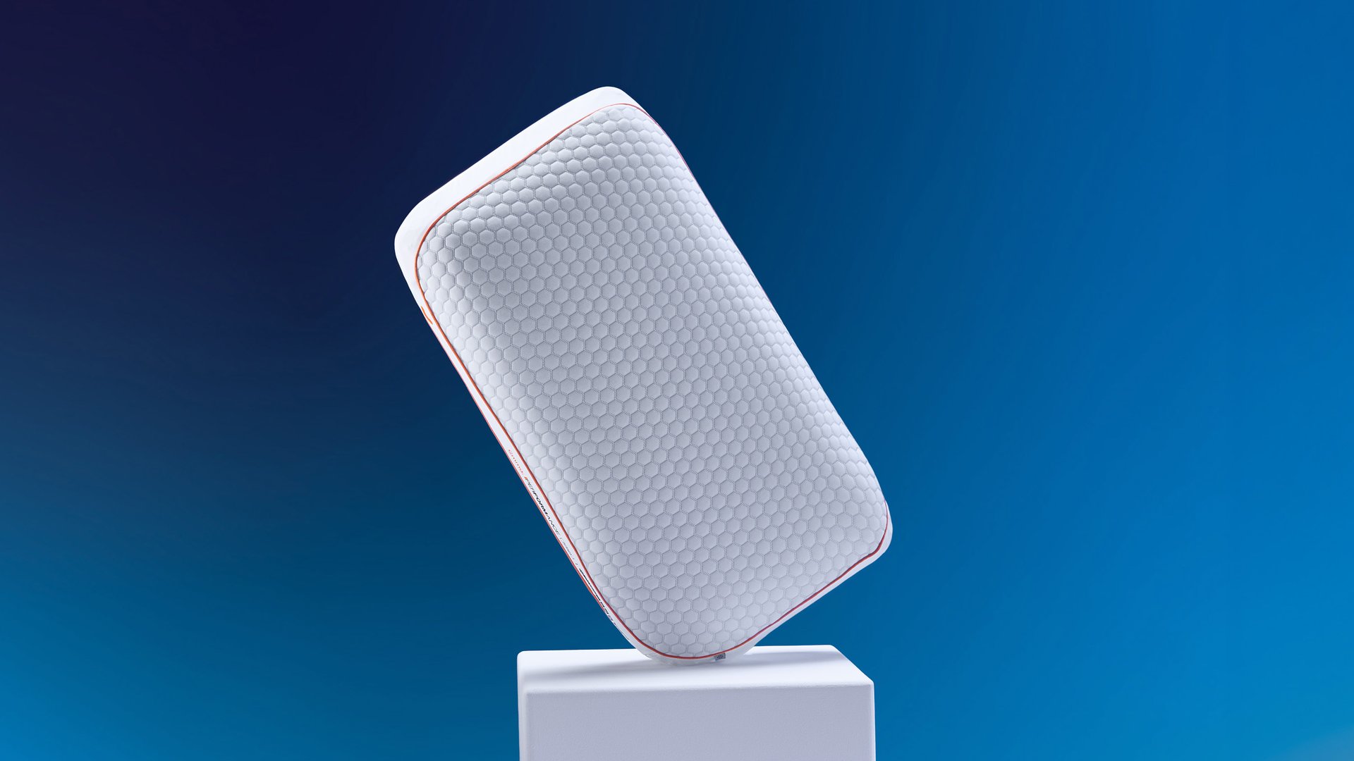 Emma Performance Adapt Pillow tilted on a white plinth showing hexagonal cover and orange trim against a blue background.