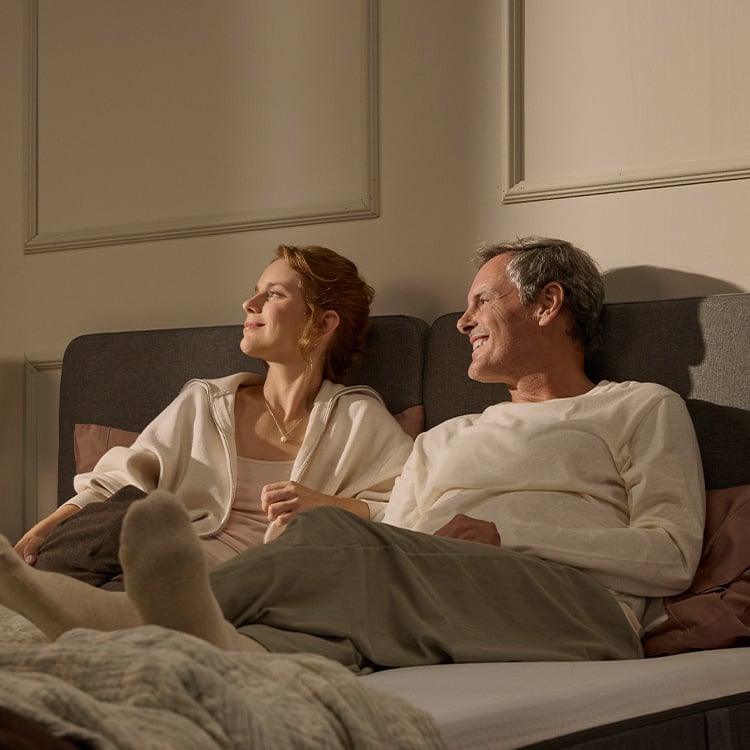 A middle-aged couple laying on an Emma Bed smiling to their right from where a beam of sunlight enters the cosy, nostalgic bedroom.