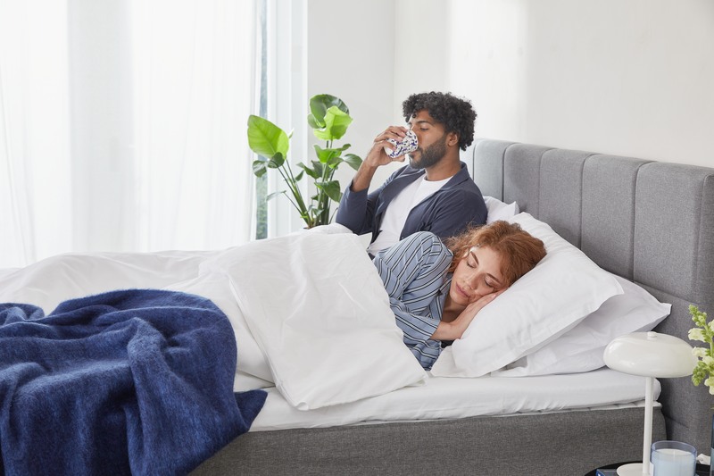 Man zit rechtop in bed en kijkt op zijn smartphone terwijl vrouw ligt onder een witte deken; grijze hoofdeinde, witte en blauwe dekens, plant en lamp op nachtkastje in lichte slaapkamer.