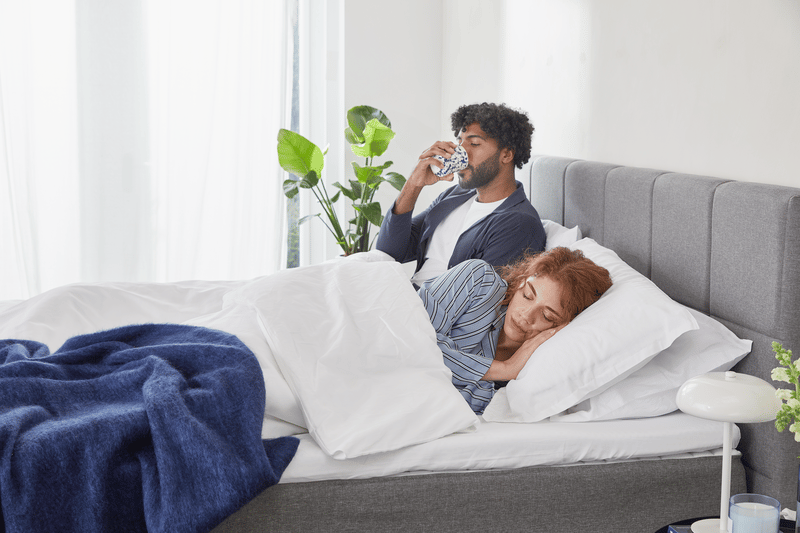 Man zit rechtop in bed en kijkt op zijn smartphone terwijl vrouw ligt onder een witte deken; grijze hoofdeinde, witte en blauwe dekens, plant en lamp op nachtkastje in lichte slaapkamer.