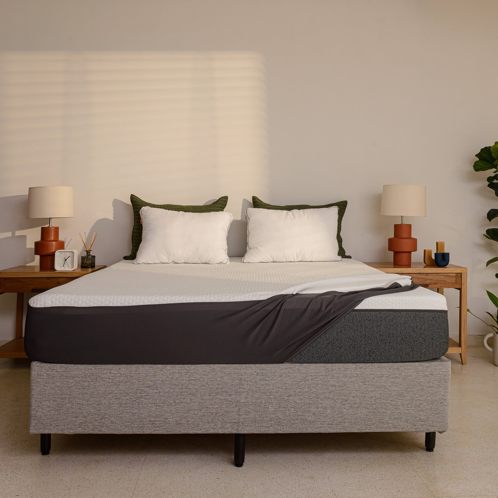 Quarto arrumado com decoração minimalista. A cama box tem sobre ela um colchão parcialmente coberto por um protetor, ou capa protetora de colchão. Ao fundo, junto à cabeceira da cama, estão travesseiros brancos. À volta da cama, duas mesas de cabeceira com candeeiros e objetos decorativos.