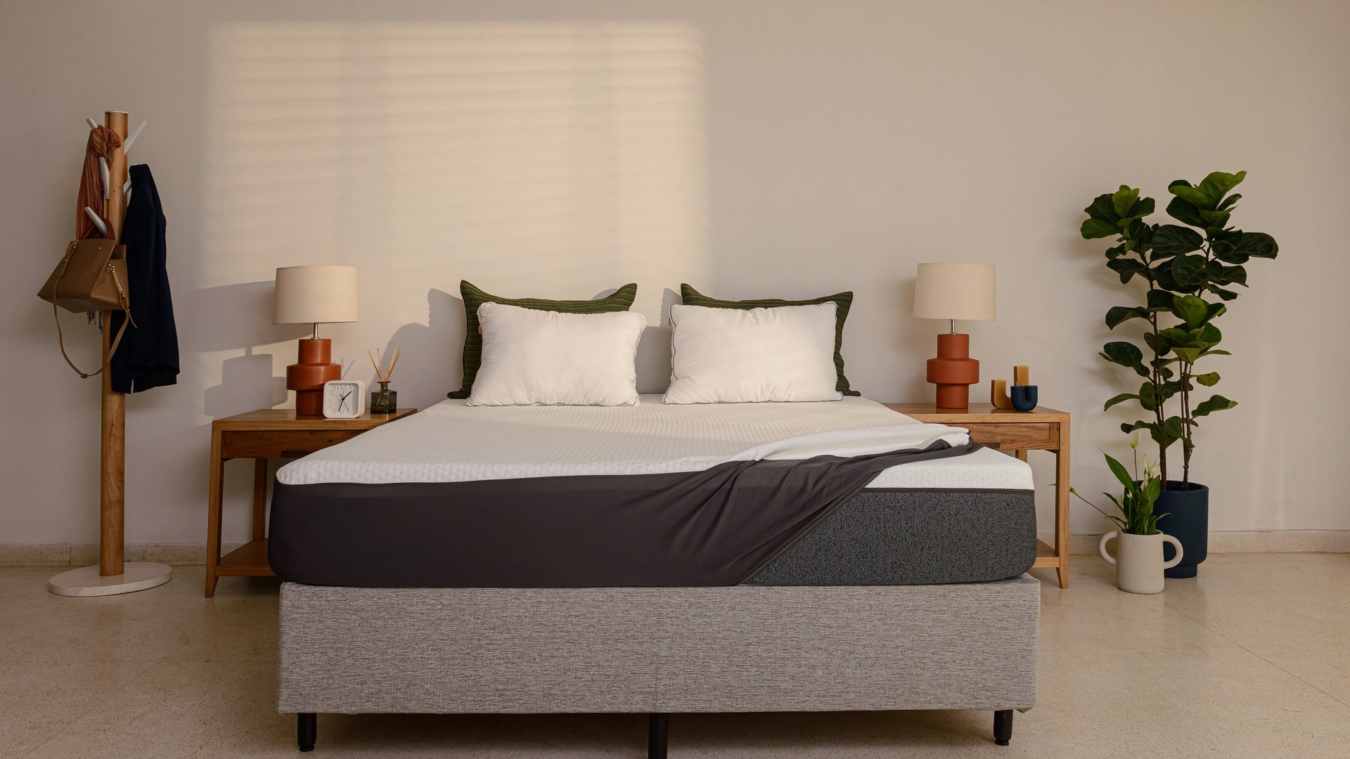 Quarto arrumado com decoração minimalista. A cama box tem sobre ela um colchão parcialmente coberto por um protetor, ou capa protetora de colchão. Ao fundo, junto à cabeceira da cama, estão travesseiros brancos. À volta da cama, duas mesas de cabeceira com candeeiros e objetos decorativos.