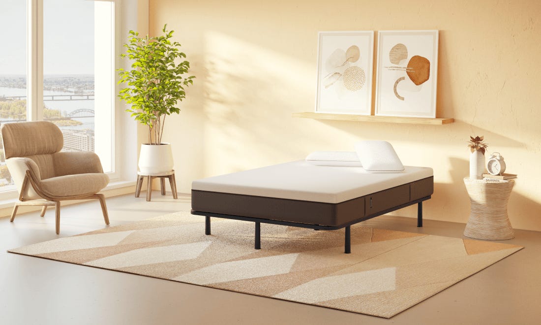 Emma Essential Bedframe met zwart metalen frame en witte matras in lichte slaapkamer—grote ramen met uitzicht op brug en bomen, beige fauteuil, groene plant en geometrisch vloerkleed creëren een moderne en rustige sfeer.