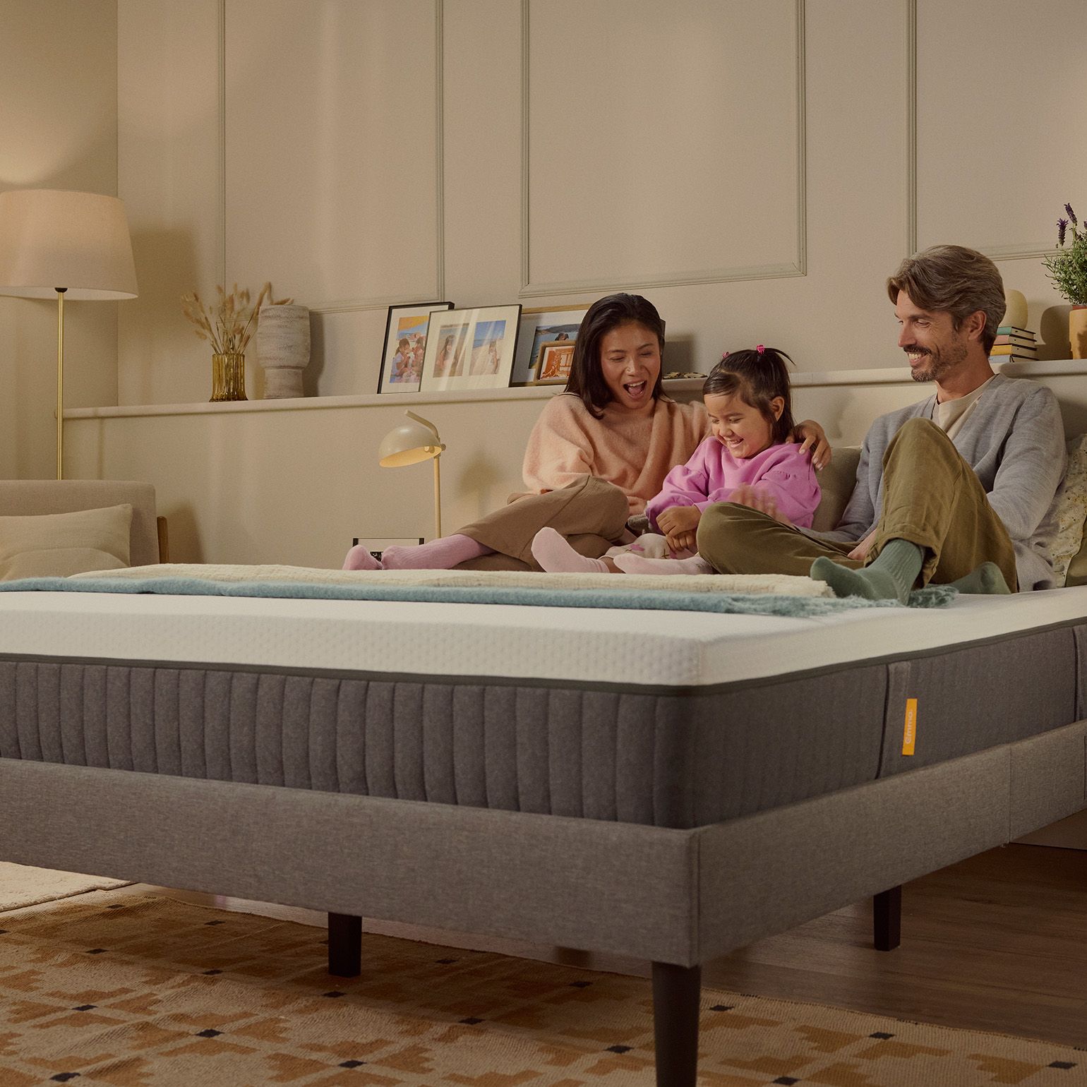 Une famille de trois personnes rit ensemble sur un matelas Emma Original dans une chambre à la lumière chaleureuse.