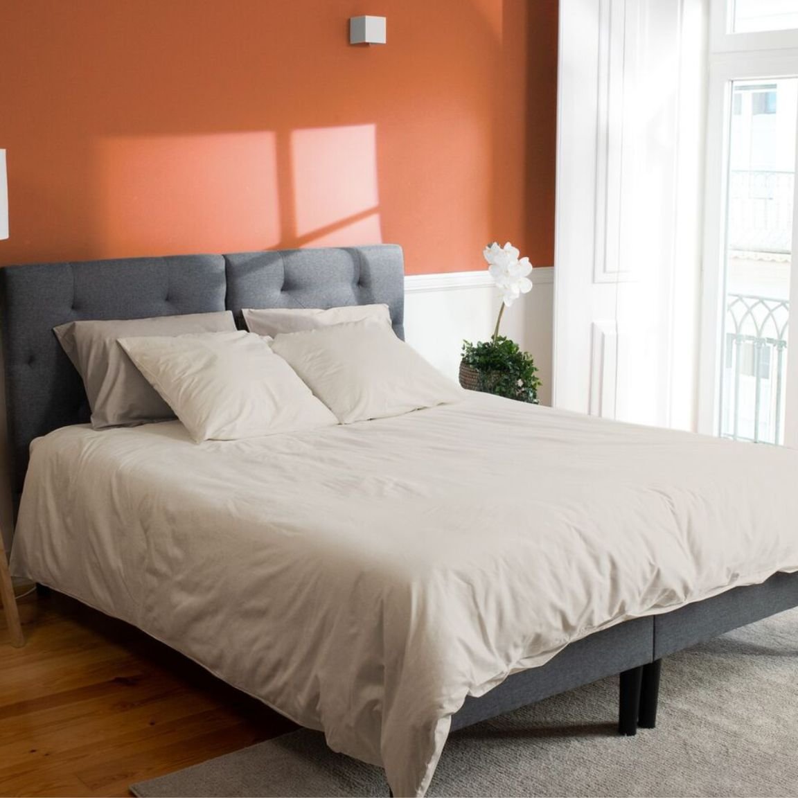 Camera da letto ordinata con parete arancione, letto con testiera grigia e decorazioni naturali.