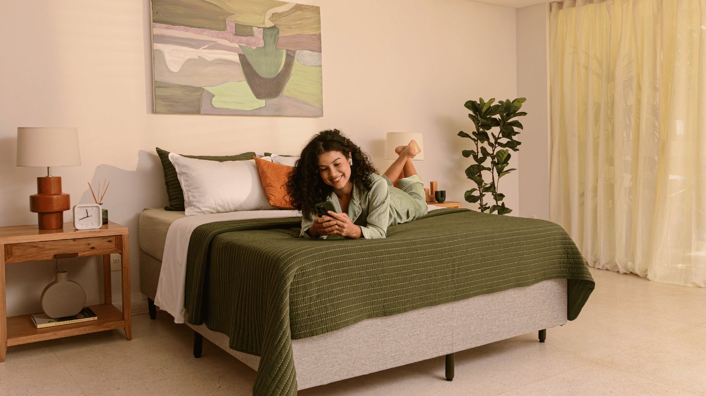 Quarto moderno e aconchegante com uma pessoa deitada de bruços sobre uma cama Emma com colcha verde, lençóis brancos Emma e almofadas coloridas. A pessoa veste roupa verde e segura um celular. O ambiente inclui uma pintura grande na parede, criado-mudo de madeira com abajur e objetos decorativos, planta em vaso ao lado da cama e cortinas claras que deixam entrar luz natural.