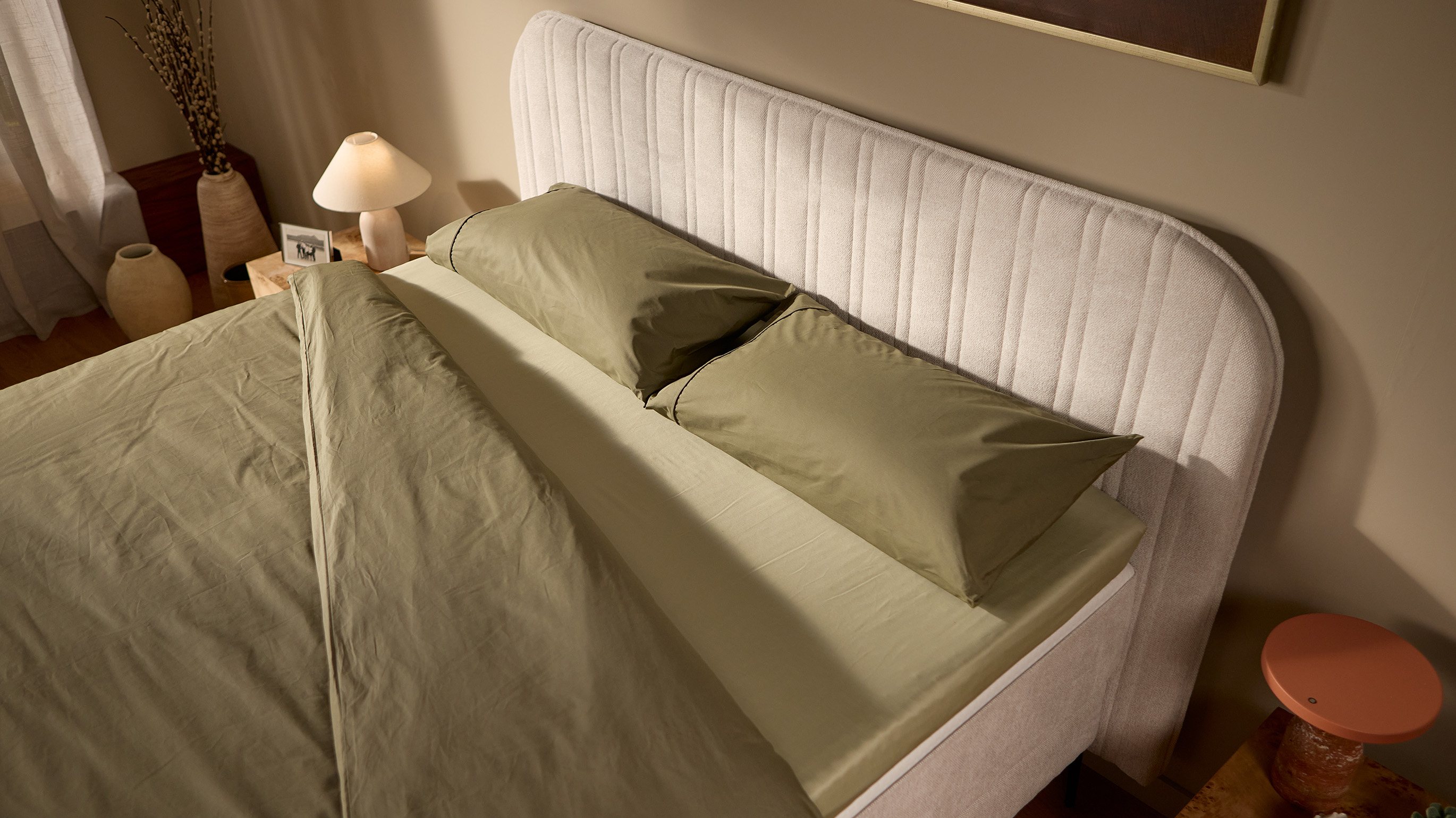 Close-up of the Emma Boxspring Bed in beige showing the curved channelled headboard with olive green pillows.
