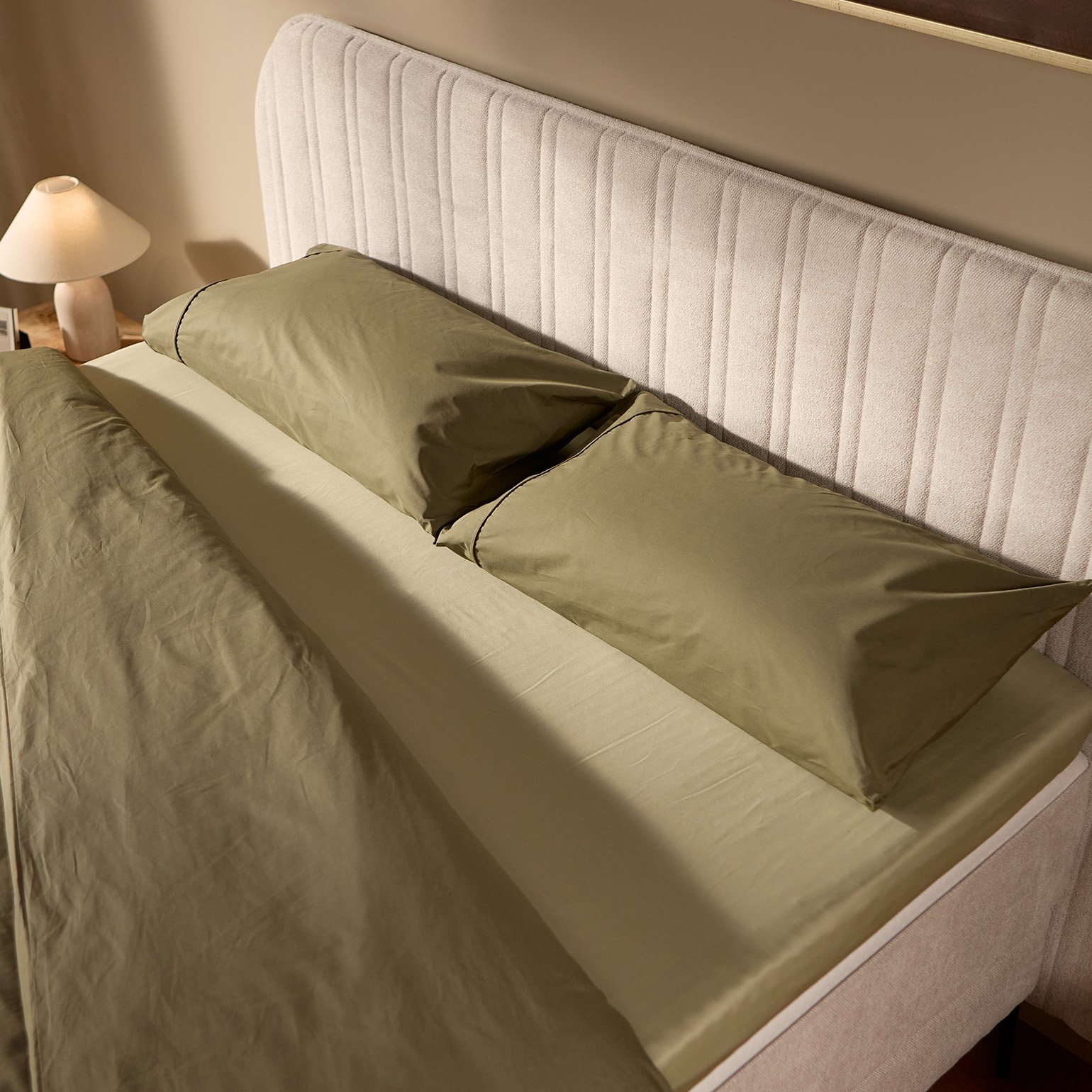Close-up of the Emma Boxspring Bed in beige showing the curved channelled headboard with olive green pillows.
