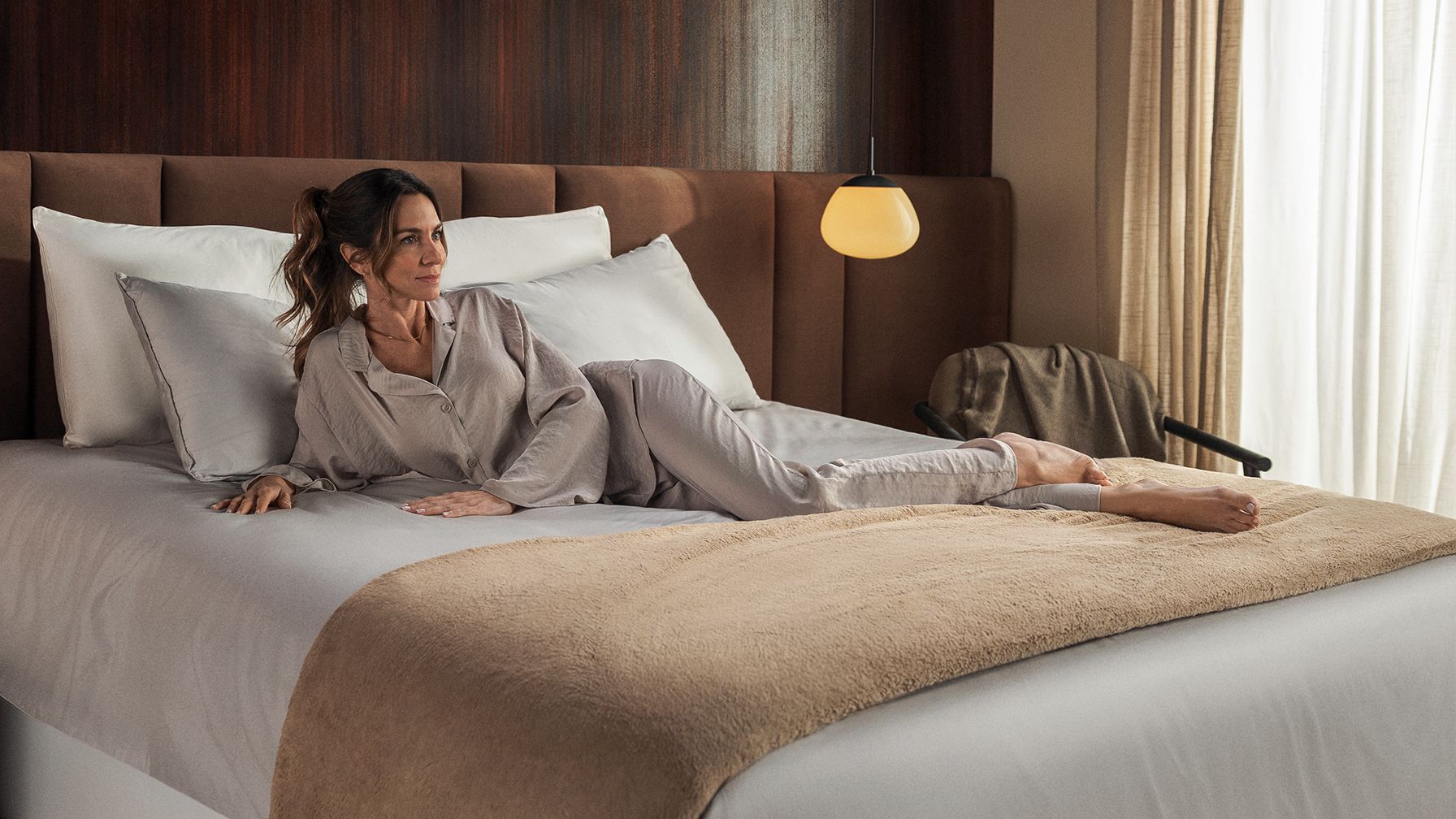 Een persoon ligt ontspannen op een opgemaakt bed met een beige plaid en witte kussens. Het bed heeft een bruin gestoffeerd hoofdbord, en de kamer is sfeervol ingericht met een gele hanglamp, beige gordijnen en een stoel op de achtergrond. De setting straalt rust en comfort uit.