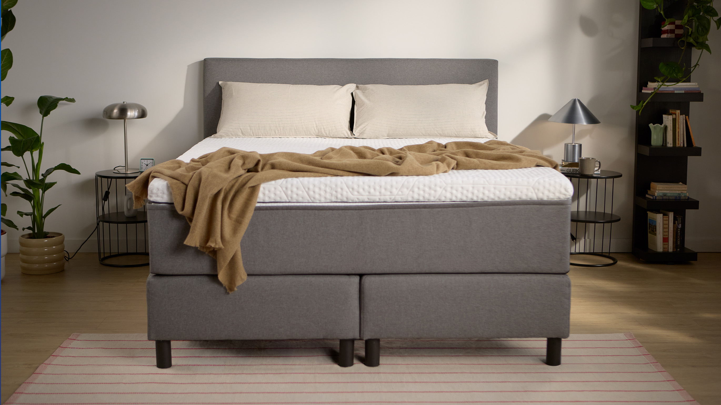 Emma Boxspring Bed in grey with upholstered headboard and dark legs, styled in a minimal bedroom with a camel throw.
