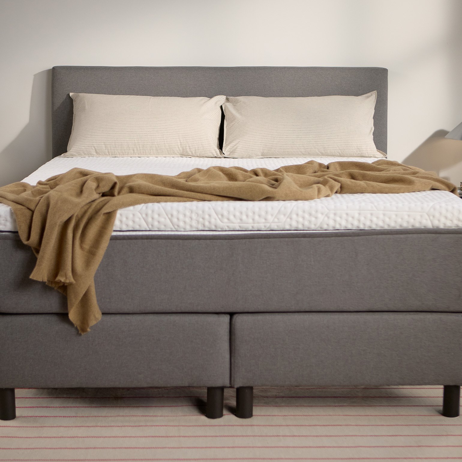 Emma Boxspring Bed in grey with upholstered headboard and dark legs, styled in a minimal bedroom with a camel throw.

