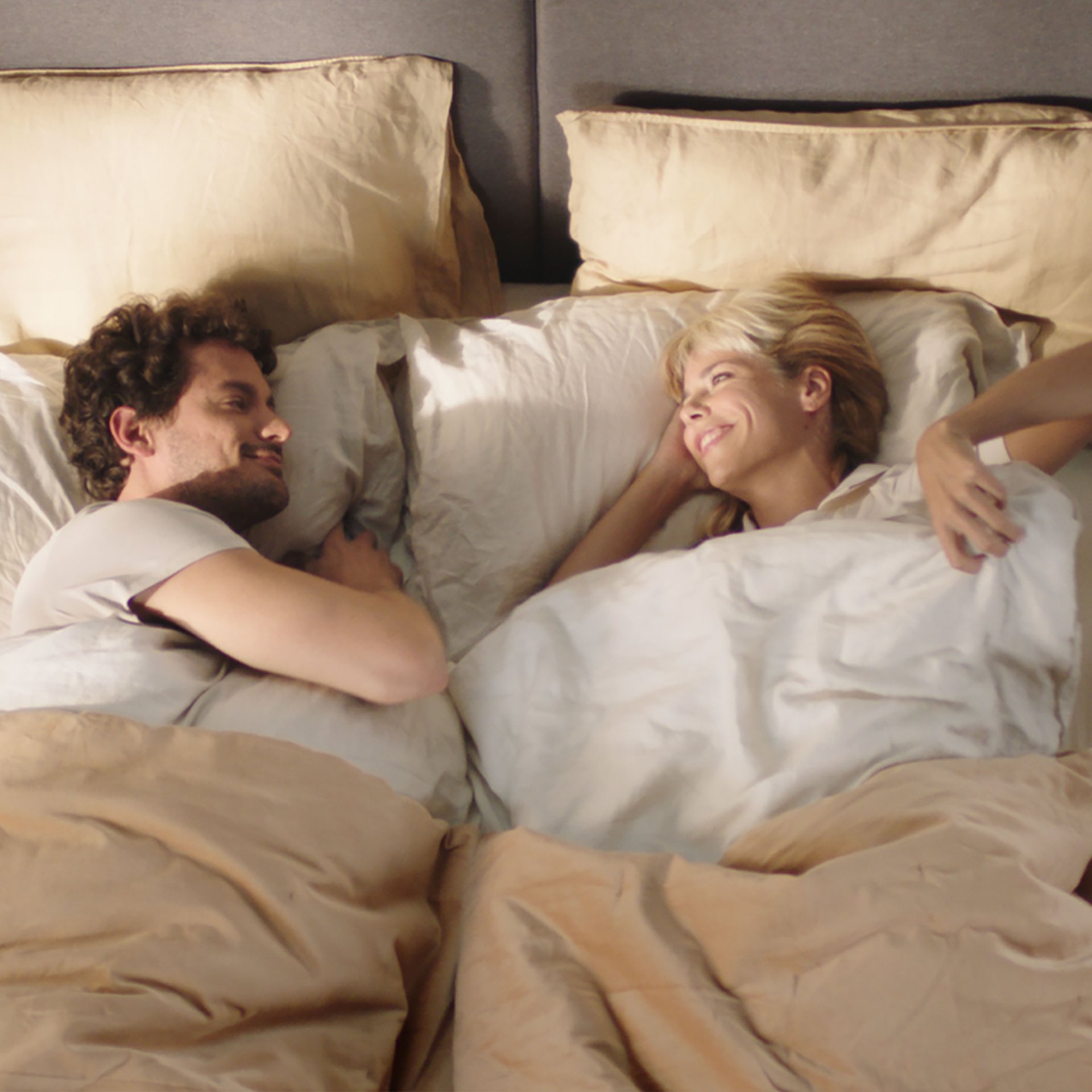 Due persone a letto con biancheria beige e bianca, libri sul lato sinistro.