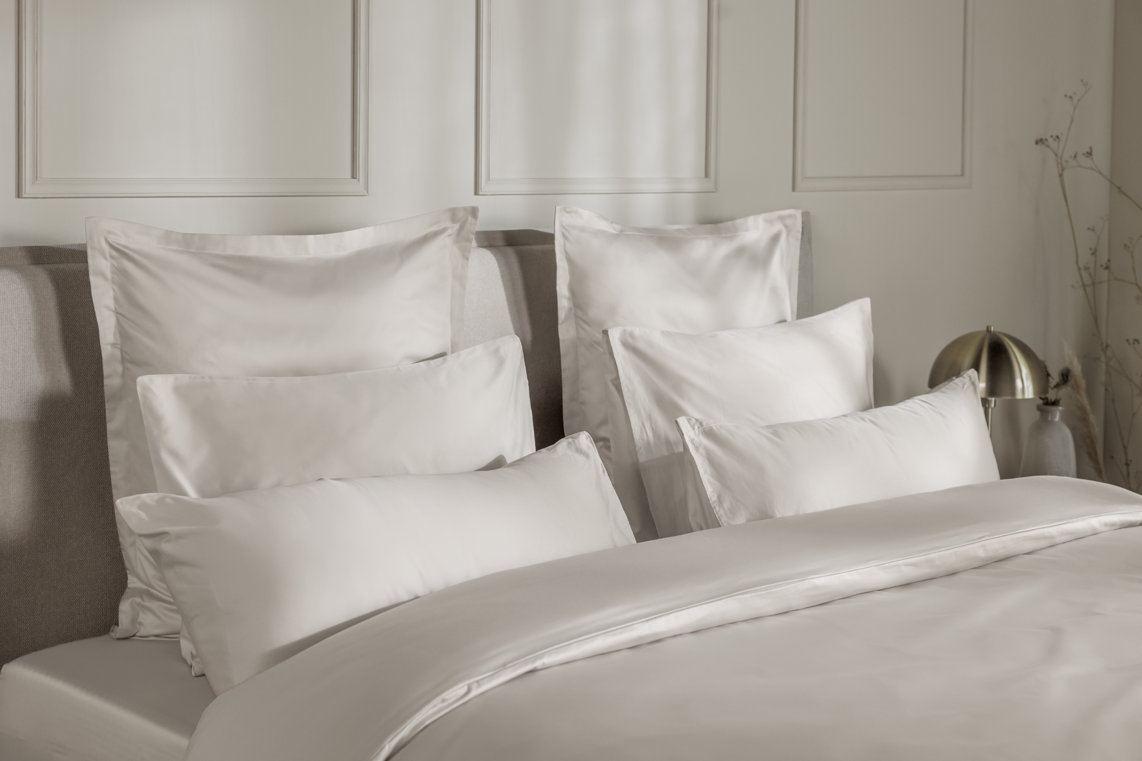 Detail van een bedhoek met een Emma Satijn Hoeslaken in een zachte beigetint, strak om een matras gespannen; hoeslaken heeft een subtiele glans en zijdezachte afwerking die het luxe karakter benadrukt; bedframe is grijs met houten poten; de setting is minimalistisch met een witte muur, links een lamp met gouden voet en rechts een vaas met droogbloemen.