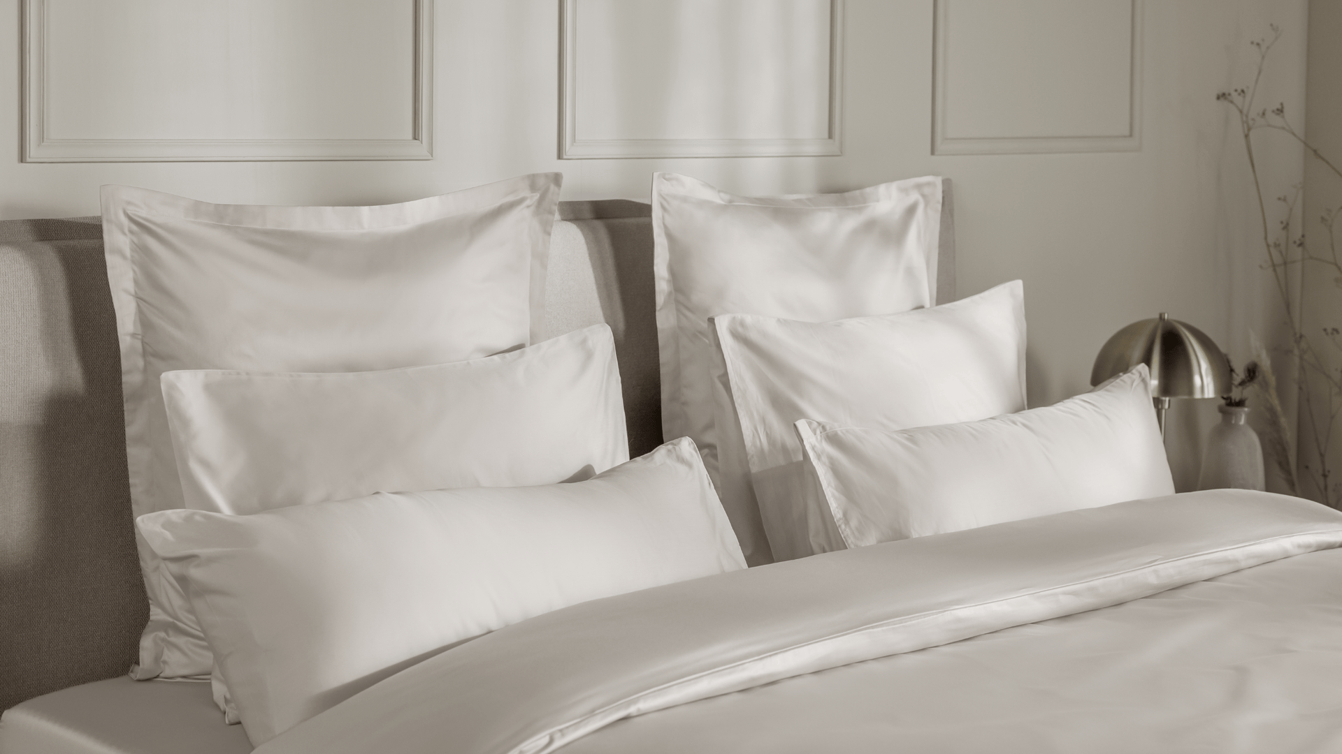 Zes witte Emma Satijnen Kussenslopen gepresenteerd op een bed: drie grote kussens achteraan en drie kleinere ervoor, allemaal voorzien van gladde, glanzende satijnstof; bed is opgemaakt met bijpassend wit dekbed; achtergrond toont een lichtgekleurde wand met sierpanelen, rechts een moderne lamp en een vaas met droogbloemen.