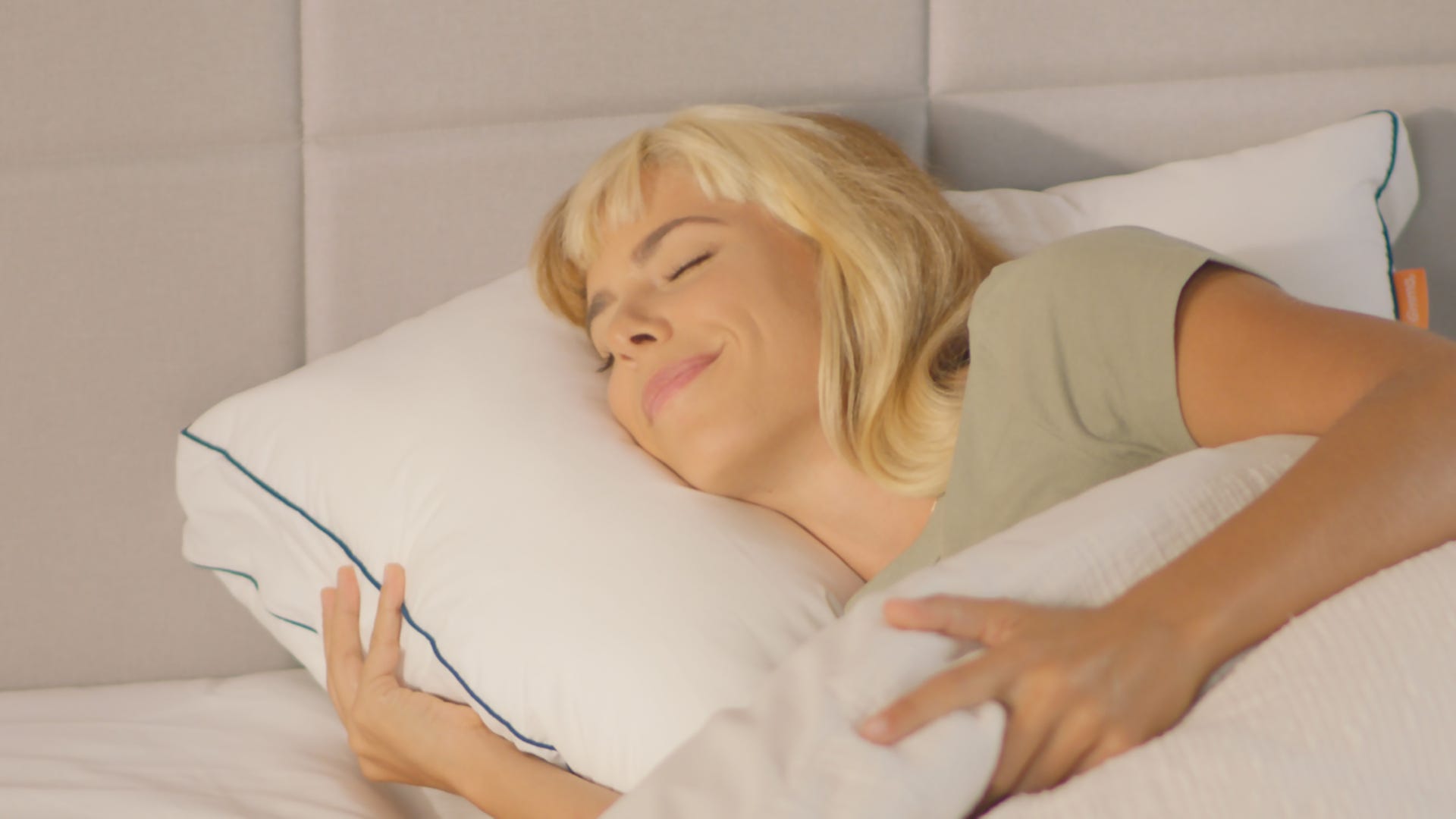 Persoon ligt ontspannen in bed met hoofd op een Emma Premium Wolkenkussen v1, wit met blauwe bies. De ogen zijn gesloten en er is een lichte glimlach zichtbaar. Het bed heeft witte lakens en een lichtgekleurde hoofdsteun, met zachte natuurlijke verlichting die een rustige en comfortabele sfeer creëert.