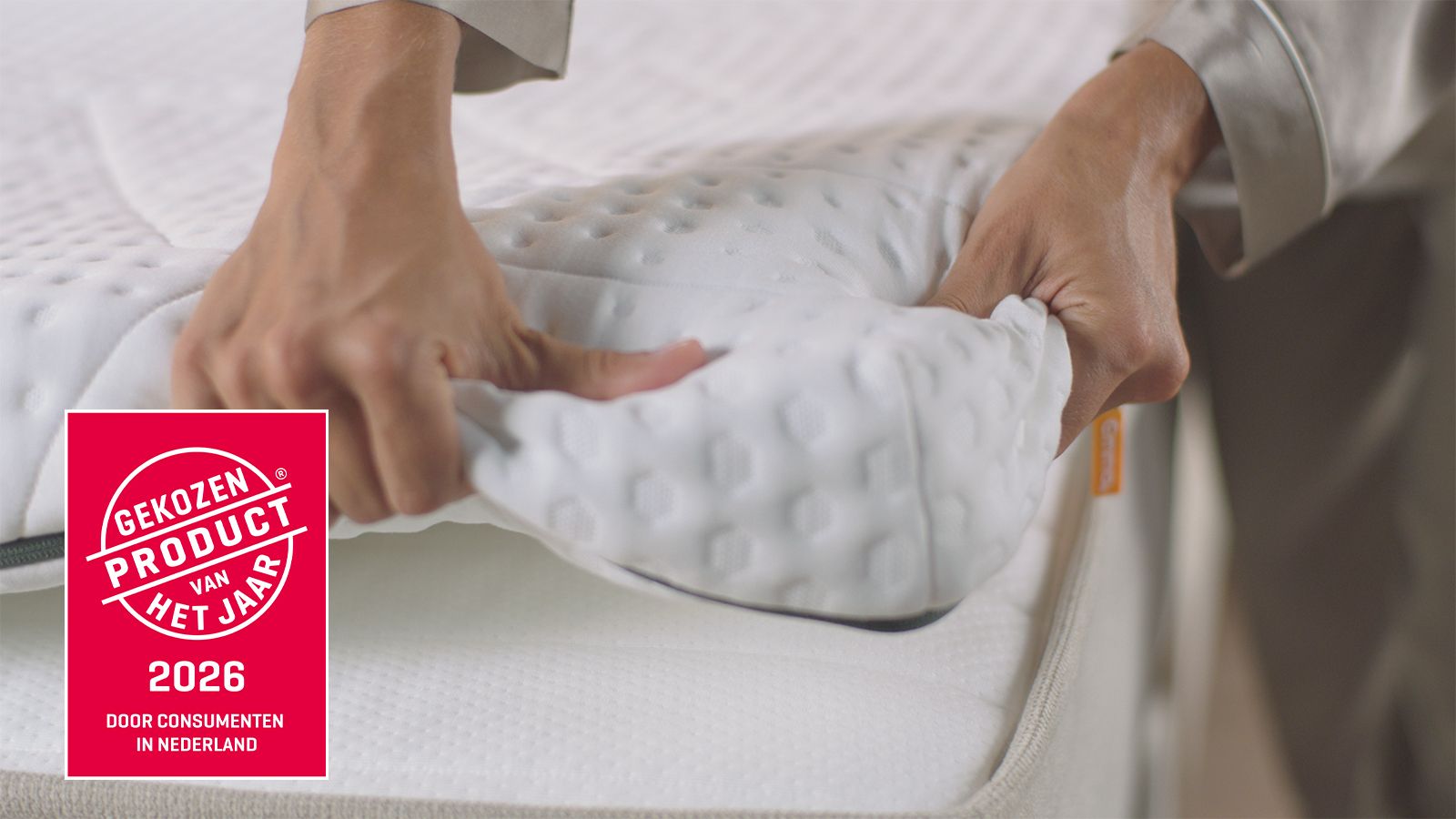 Handen tillen de hoek van een matrasbeschermer op om de structuur van het matras te tonen; links staat een rood keurmerk met ‘Gekozen Product van het Jaar 2026 – Door consumenten in Nederland’.