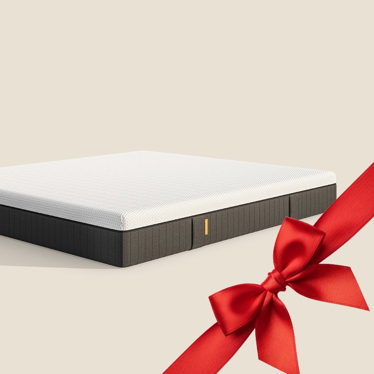 Lit moderne avec tête de lit grise et pieds noirs, recouvert de draps blancs. Deux oreillers blancs et une pile de linge plié sont posés sur le matelas. Arrière-plan beige avec une forme orange abstraite.