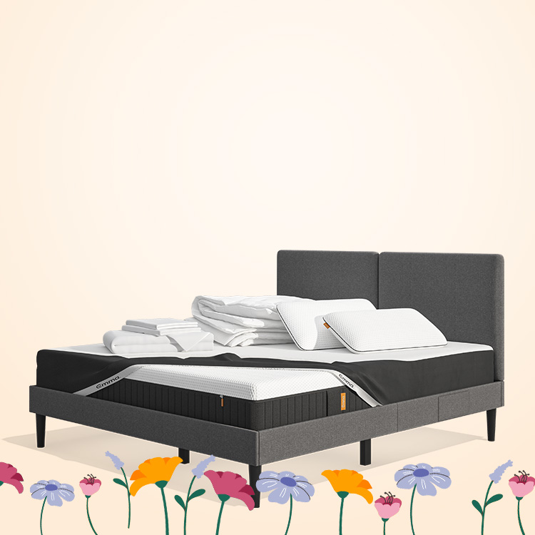 Lit moderne avec tête de lit grise et pieds noirs, recouvert de draps blancs. Deux oreillers blancs et une pile de linge plié sont posés sur le matelas. Arrière-plan beige avec une forme orange abstraite.