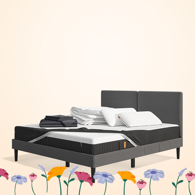 Lit moderne avec tête de lit grise et pieds noirs, recouvert de draps blancs. Deux oreillers blancs et une pile de linge plié sont posés sur le matelas. Arrière-plan beige avec une forme orange abstraite.