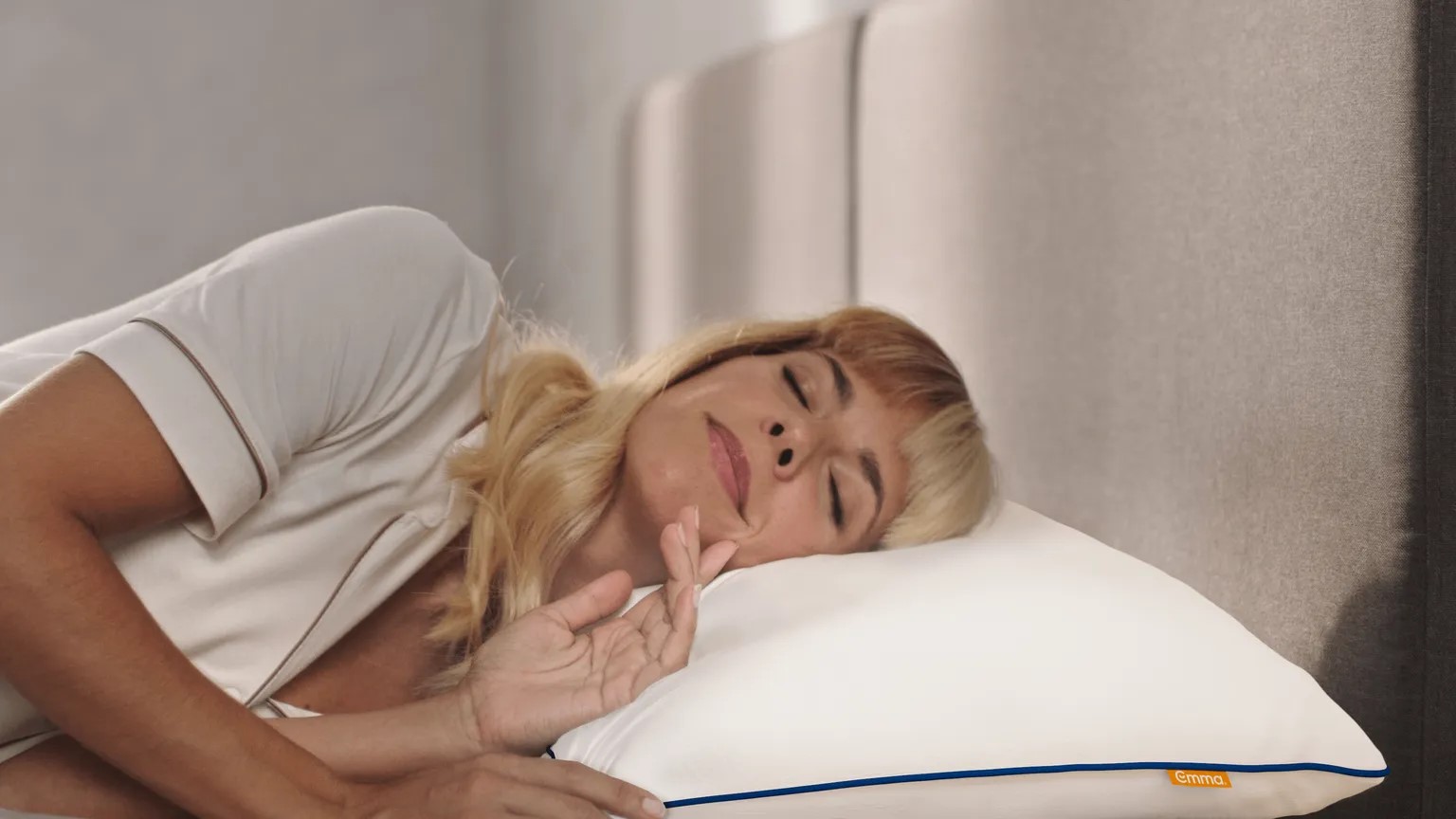 Persoon ligt op een bed en rust het hoofd op een wit Emma Wolkenkussen met blauwe bies en oranje merklabel; witte kleding en grijs hoofdeinde vormen een rustgevende setting.