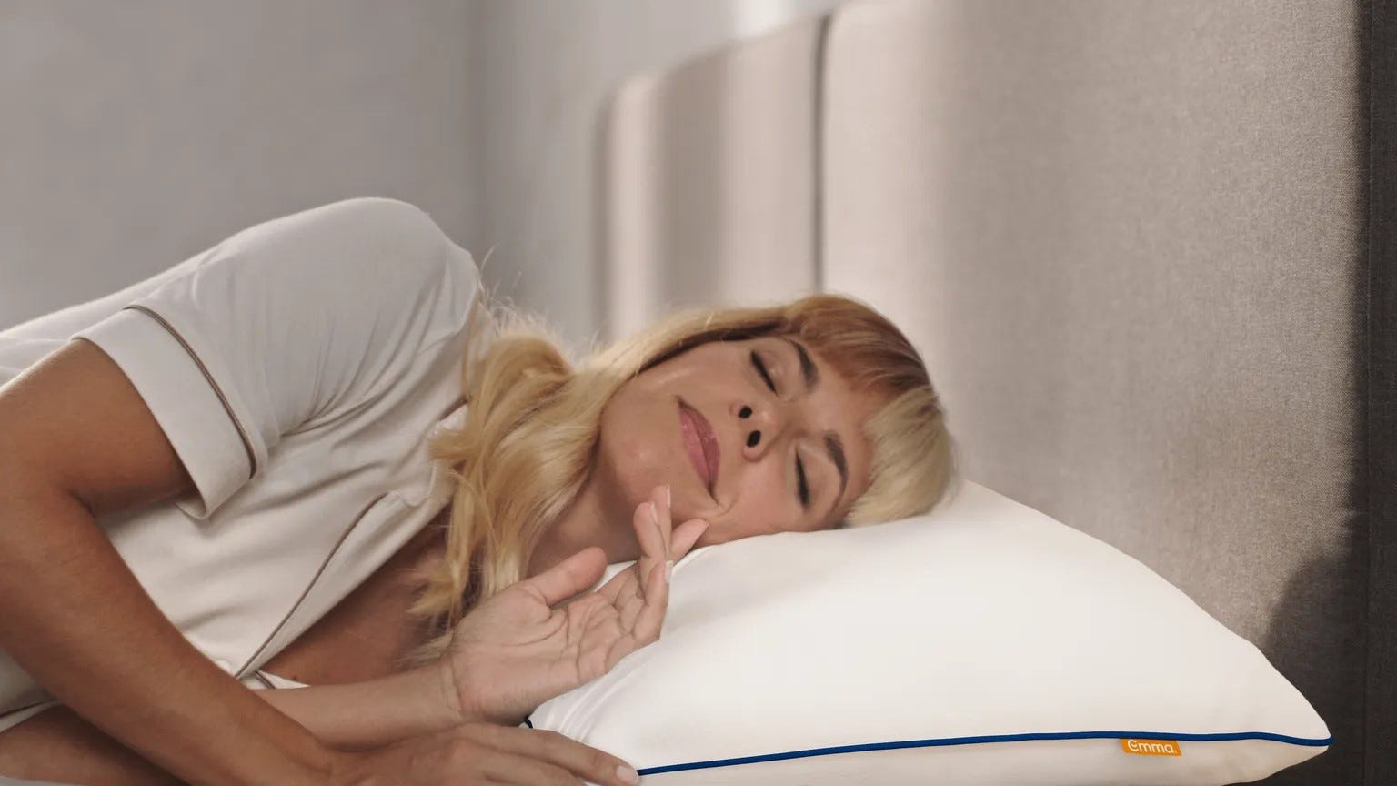 Persoon ligt op een bed en rust het hoofd op een wit Emma Wolkenkussen met blauwe bies en oranje merklabel; witte kleding en grijs hoofdeinde vormen een rustgevende setting.