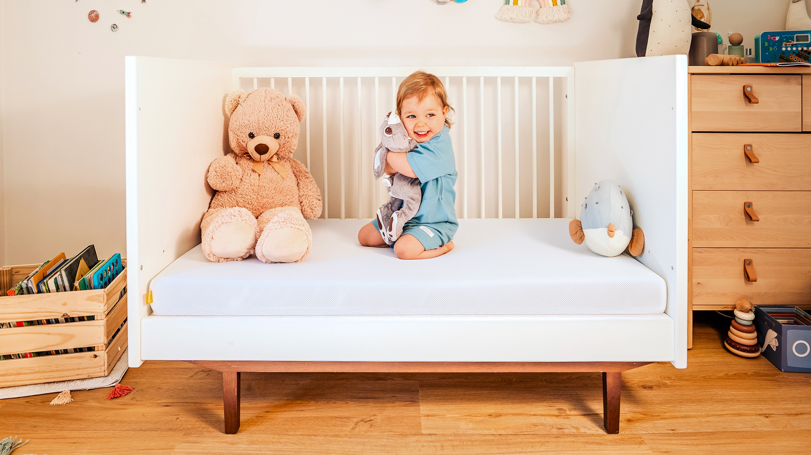 Kleinkind sitzt lachend im weißen Gitterbett mit Stofftieren auf einer Matratze.
