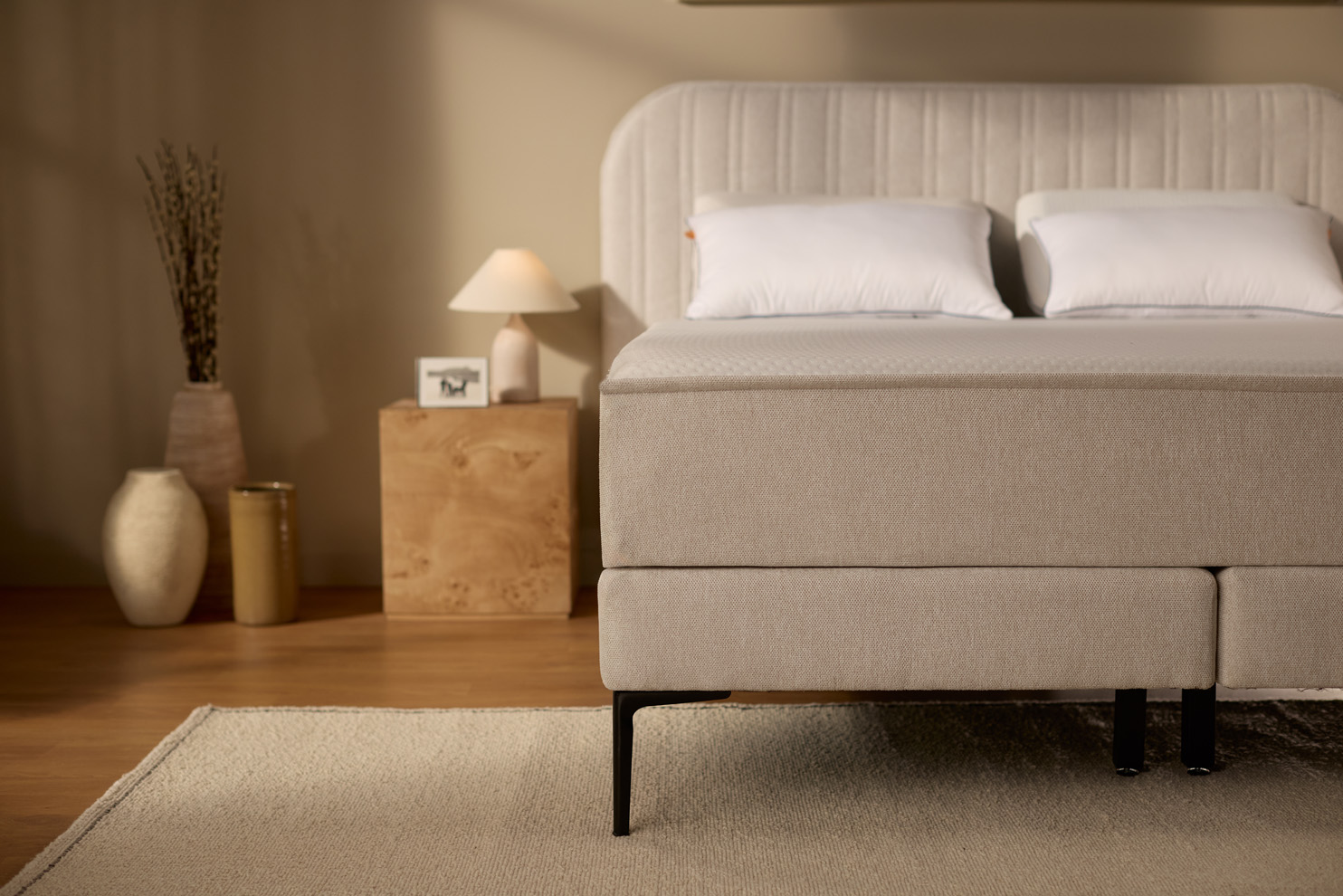 Close‑up van de lichtbeige gestoffeerde boxspringbasis met stoffen afwerking en ondersteuningspoten, getoond in een slaapkamer met kussens tegen het hoofdbord.