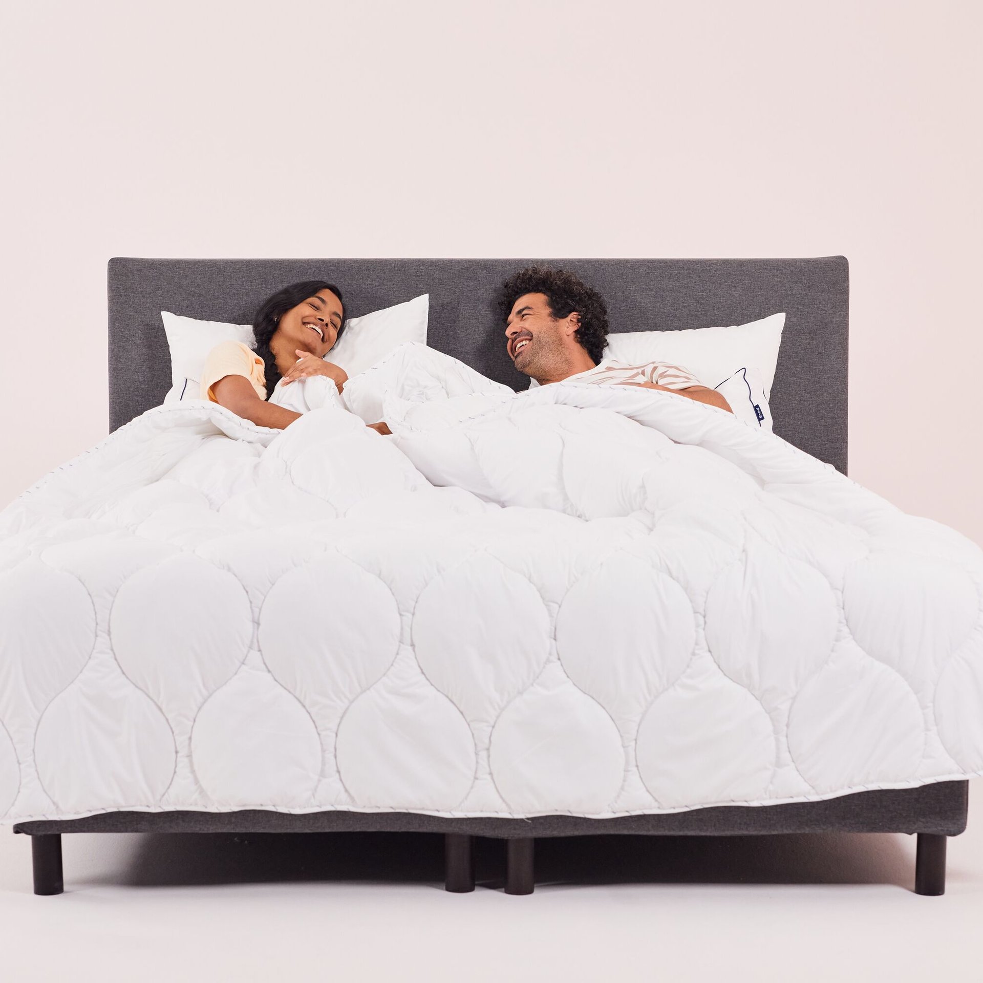 Due persone a letto sotto un piumone bianco, con testiera grigio scuro e sfondo rosa chiaro.