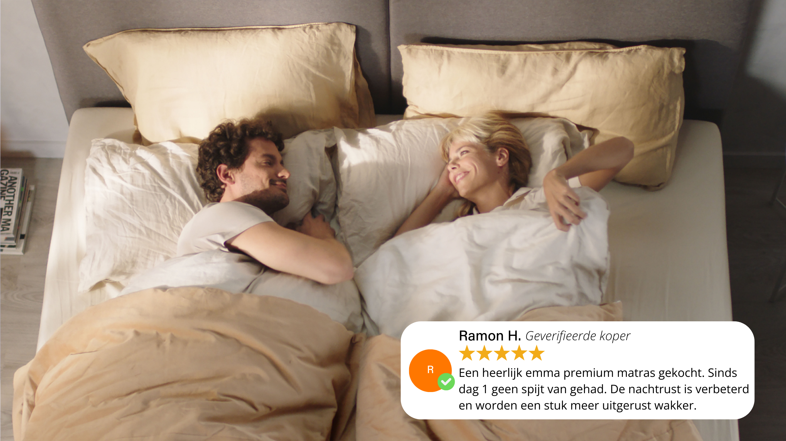 Twee personen liggen ontspannen onder beige dekens op een bed met bijpassende kussens; links een stapel boeken op het nachtkastje, rechtsonder een klantbeoordeling met vijf sterren waarin het Emma Premium matras wordt geprezen.