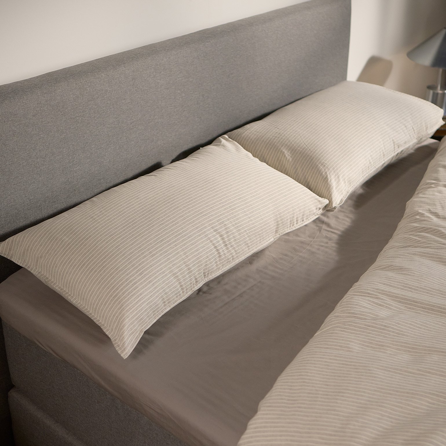 Close-up of the Emma Boxspring Bed in grey showing the upholstered headboard with cream striped pillows.
