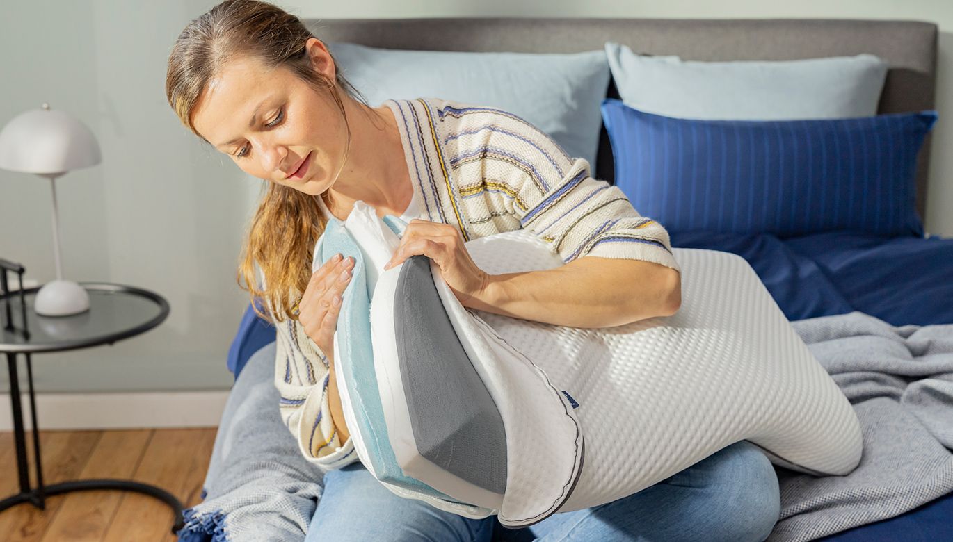 Une femme assise sur un lit ouvre la housse d'un grand oreiller blanc et en inspecte l'intérieur. Le lit est recouvert d'une couverture bleue et d'oreillers, et une petite table ronde avec une lampe se trouve à côté d'elle.