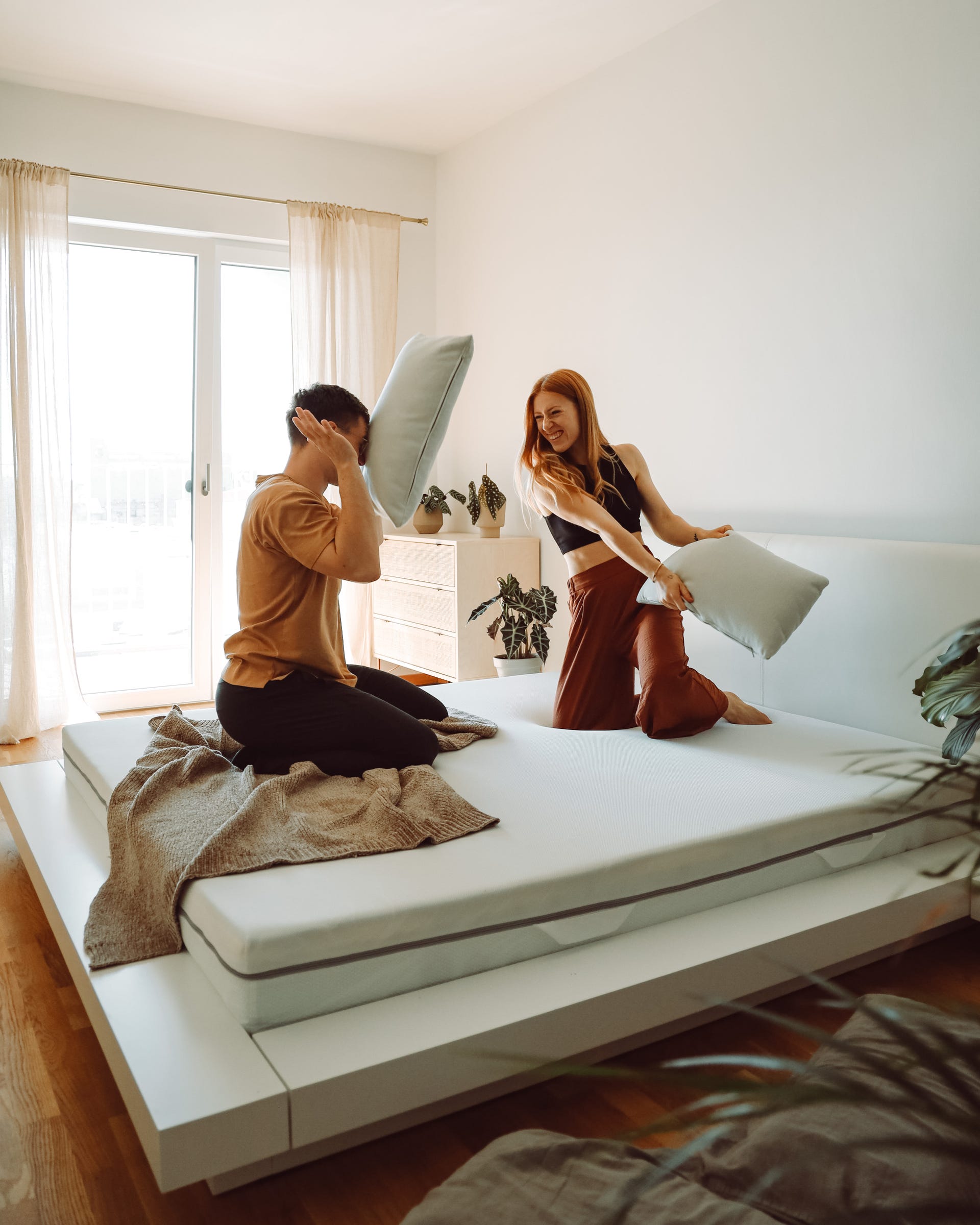 Paar macht Kissenschlacht auf einem weißen Bett mit Emma Matratze in heller Schlafzimmerumgebung.
