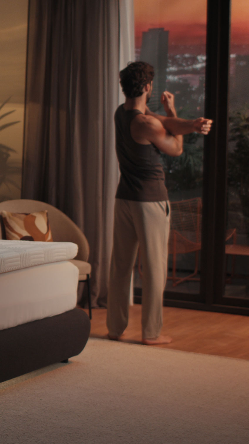 Man stretching by bedroom window at sunrise with Emma Performance Topper on bed in background.