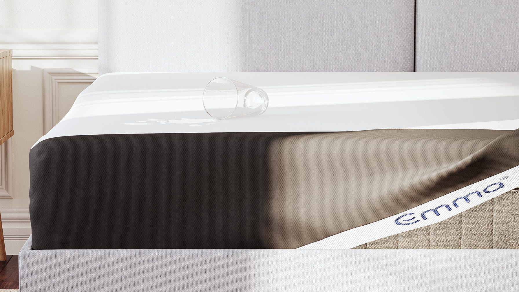 Emma matras met witte matrasbeschermer Emma-logo, geplaatst op een houten bedframe met decoratieve witte wandpanelen op de achtergrond; een omgevallen drinkglas ligt op de matras, naast een houten nachtkastje.