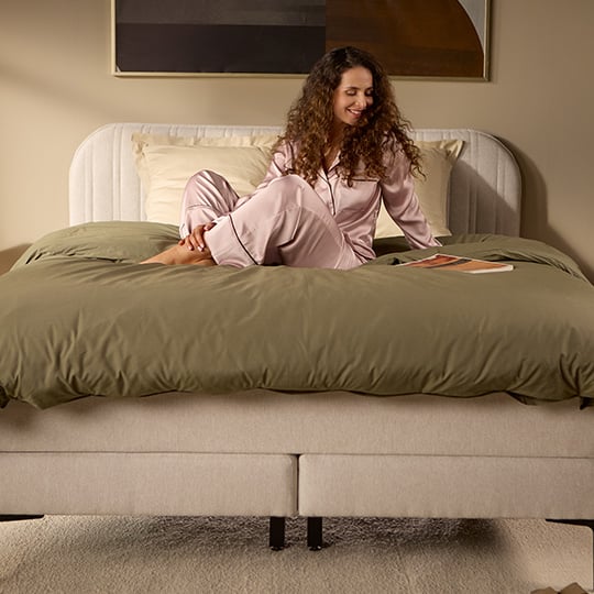 Beige upholstered bed with white mattress and green duvet, shown with a seated person.