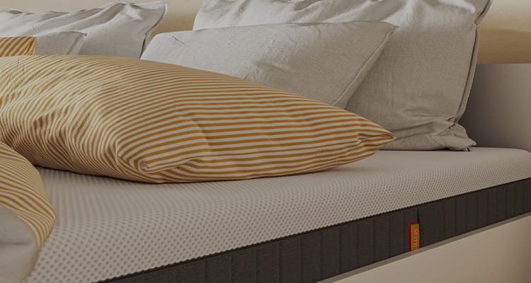 Close-up van een bed met een witte, textuurmatras met zwarte zijpanelen en een oranje label met het Emma-logo. Op het bed liggen meerdere kussens, waaronder een geel-wit gestreept kussen en lichtgrijze kussens. De afbeelding benadrukt het comfort en de hoogwaardige afwerking van het bed.