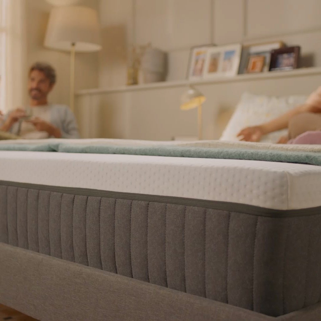 Person sitting on a bed with a white mattress in a bright bedroom.
