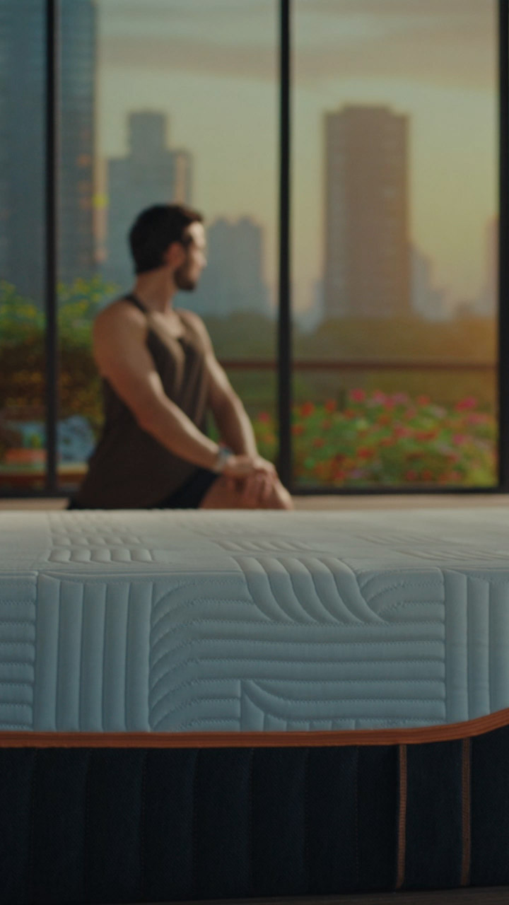 Side view of an Emma mattress with textured top and orange trim; a man in workout clothes stretches near a window with a sunrise city view.