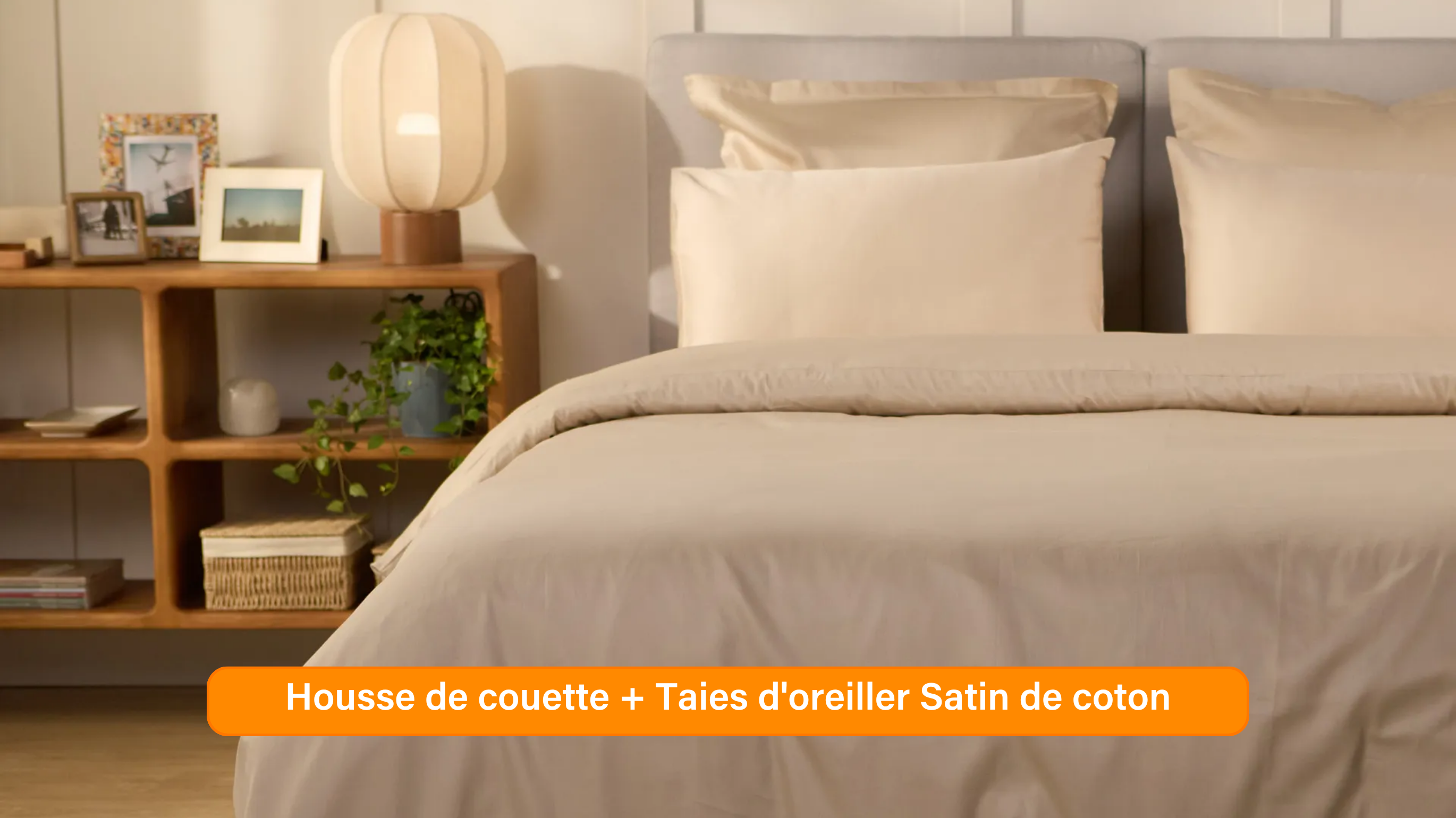 Chambre élégante avec lit soigneusement préparé, habillé d’une parure de lit 100 % satin de coton de couleur blanche ou beige clair. Plusieurs oreillers sont disposés contre un mur à panneaux. De chaque côté du lit, des tables de chevet avec lampe et objets décoratifs. Un banc rose est placé au pied du lit, assorti à un tabouret près de la table de chevet. Le sol en bois clair et les murs neutres créent une ambiance douce et raffinée.