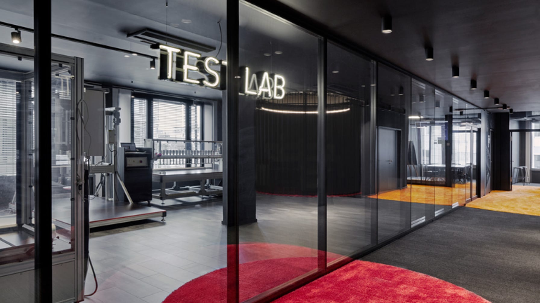 Moderne testlabruimte met glazen wanden, verlichte "TEST LAB"-letters, industriële inrichting en testapparatuur; kleurrijke ronde vloerkleden en veel licht creëren een high-tech sfeer.