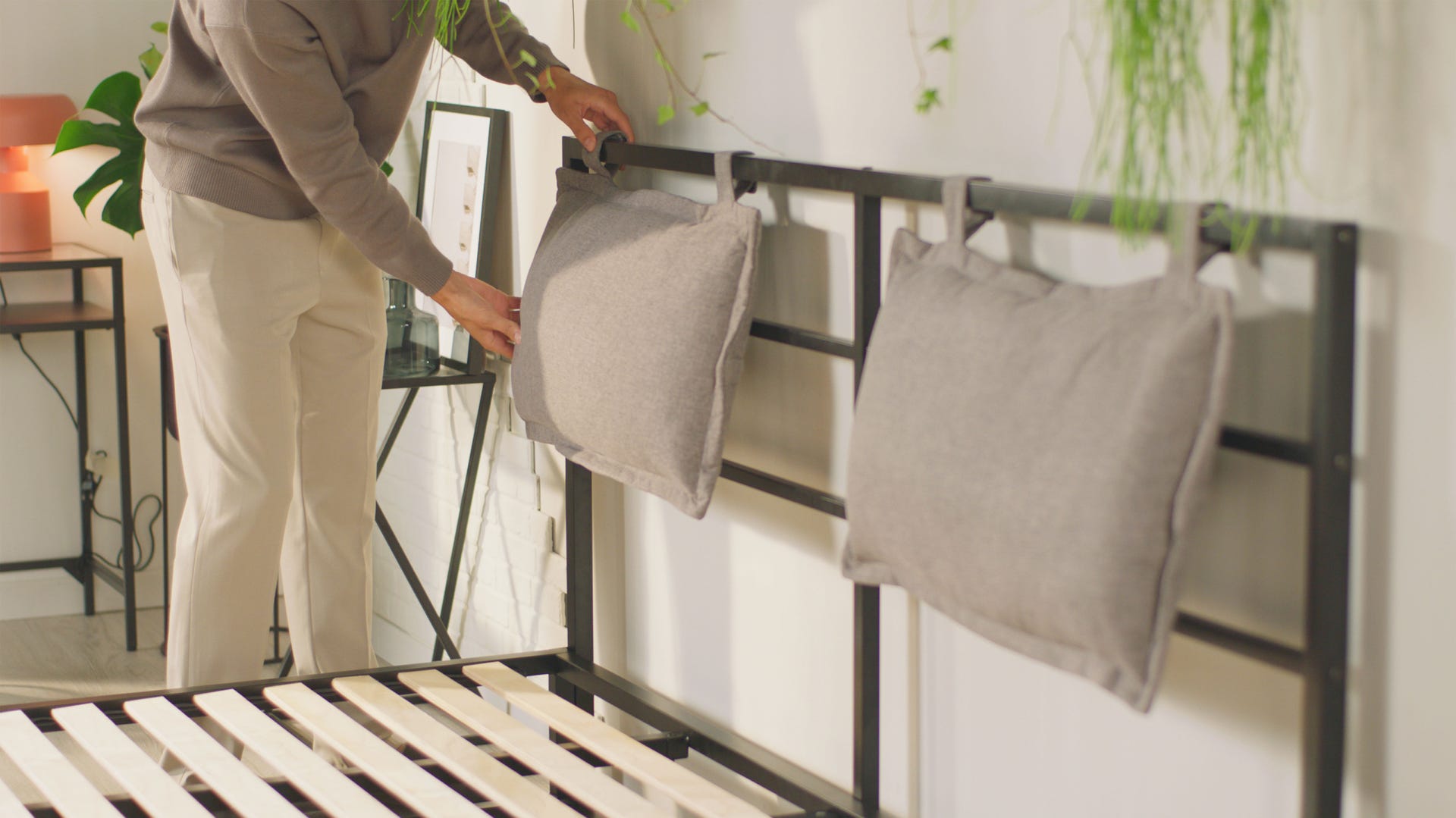 Personne fixant deux coussins gris sur la tête de lit en métal noir à l’aide de boucles en tissu. Le lit est équipé de lattes en bois visibles, sans matelas. À droite, petite table avec lampe et plante suspendue. À gauche, table d’appoint et éléments décoratifs. Ambiance moderne et minimaliste, avec touches naturelles.