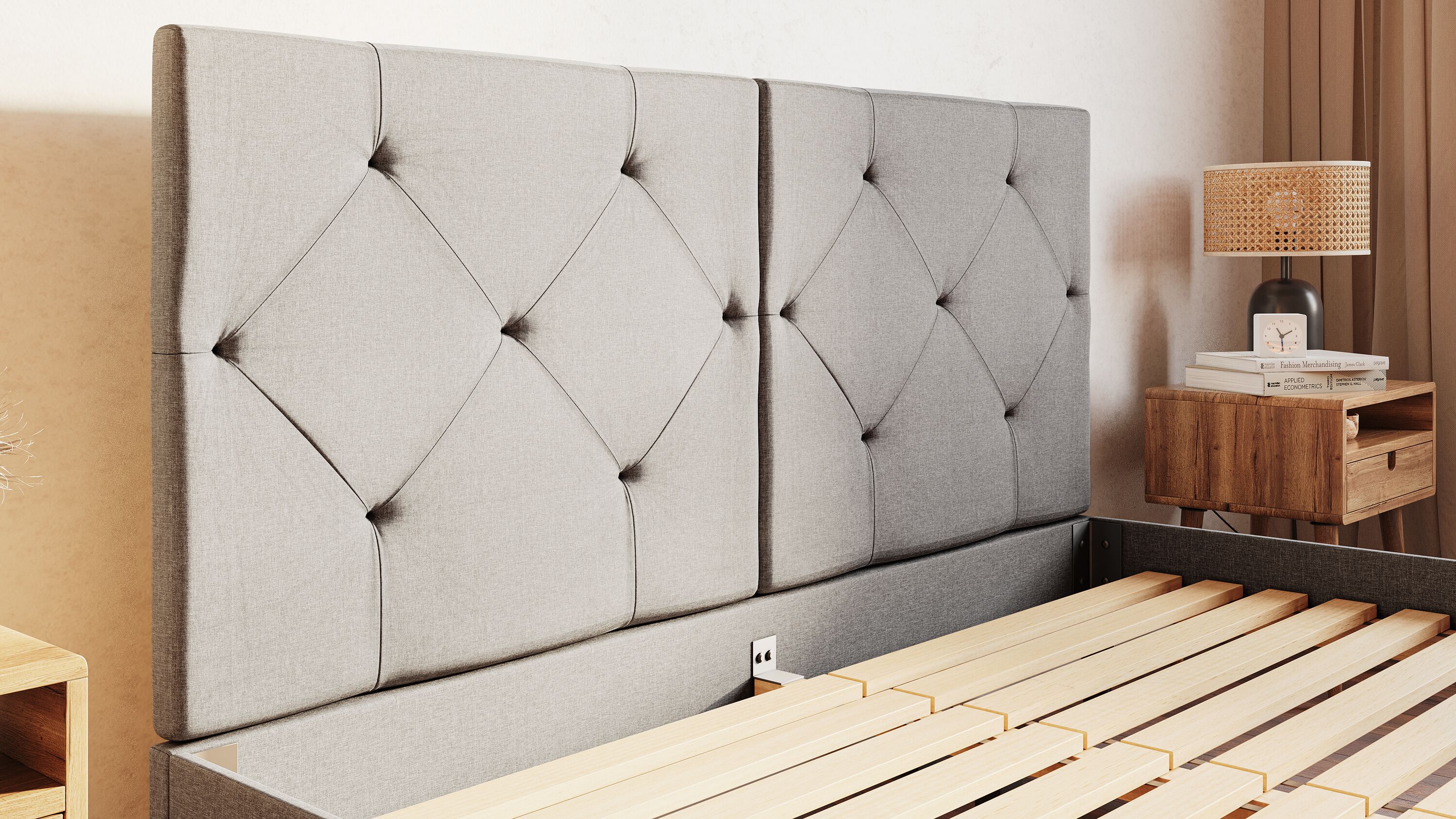 Emma Original Bed met lichtgrijs gecapitonneerd hoofdbord en gestoffeerd frame; houten lattenbodem zichtbaar zonder matras; bed staat op beige tapijt tegen crèmekleurige muur; rechts een houten nachtkastje met lamp, boeken en klok; rustige, minimalistisch ingerichte slaapkamer.