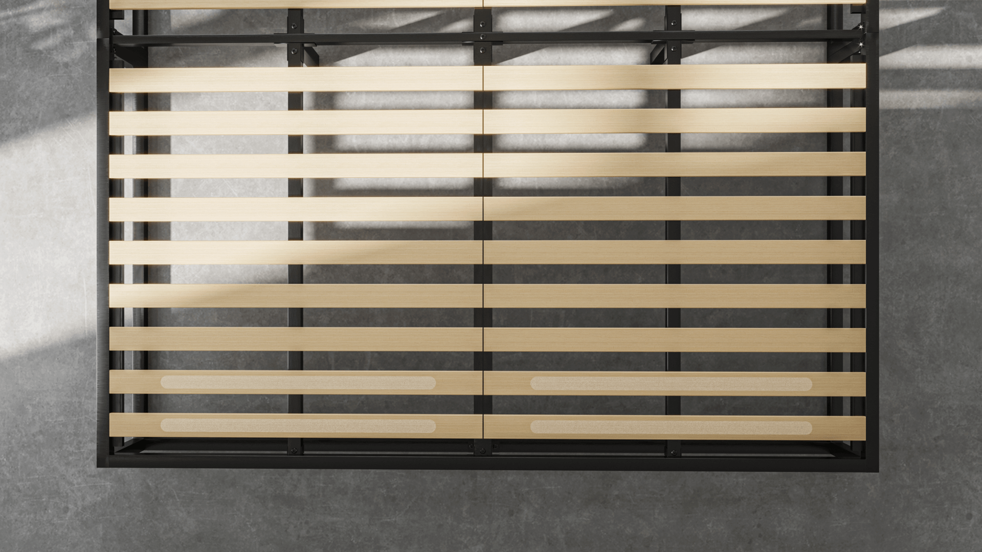 Zwart metalen bedframe met houten lattenbodem, geplaatst op grijze betonnen vloer; latten gelijkmatig verdeeld, twee onderste latten met extra versterkingspatches; lichtinval van links werpt schaduwen op vloer en bedframe.