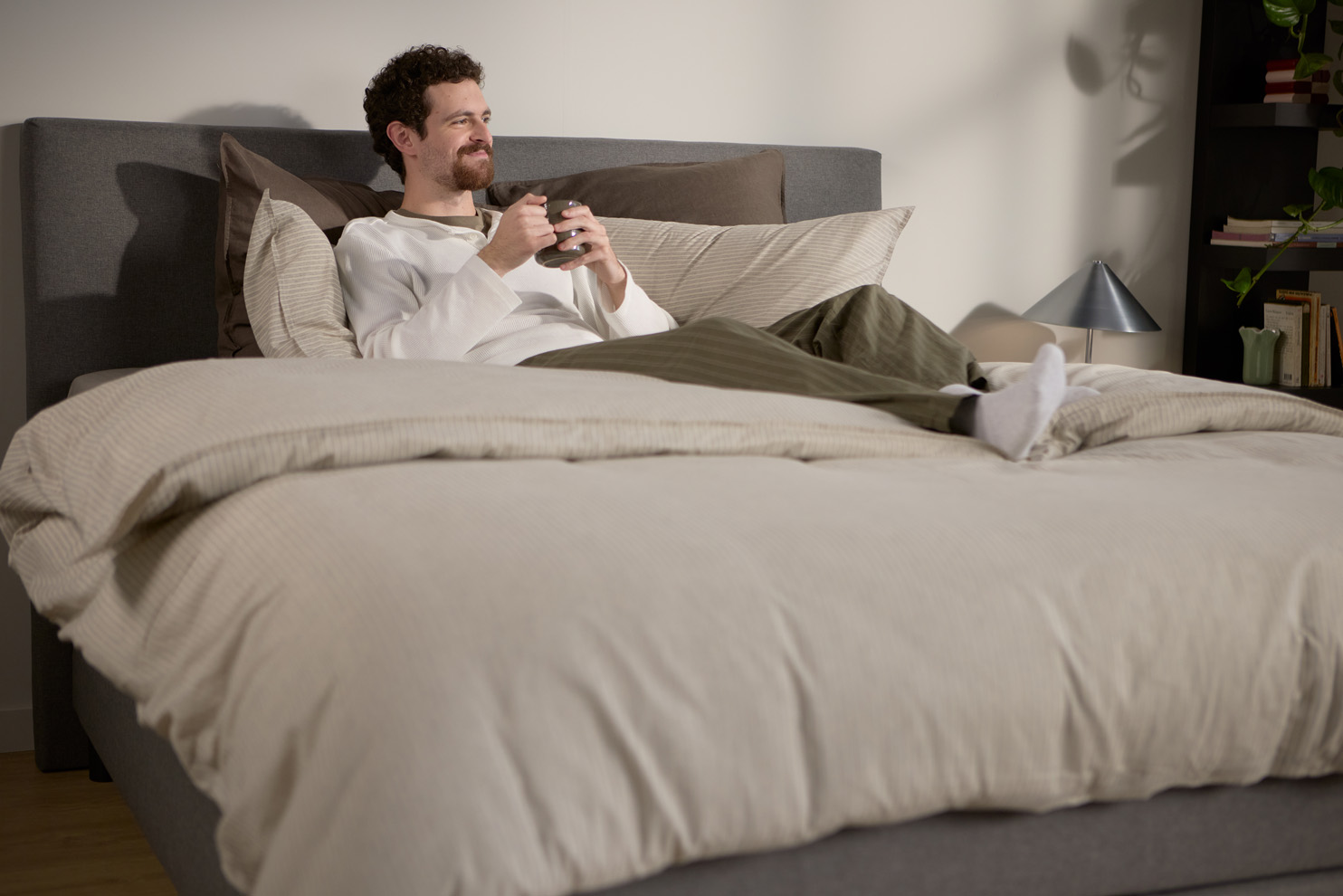 Iemand zit op een bed met een zacht, gestoffeerd hoofdbord, meerdere laagjes kussens en een dekbed in neutrale tinten, in een zacht verlichte slaapkamer.