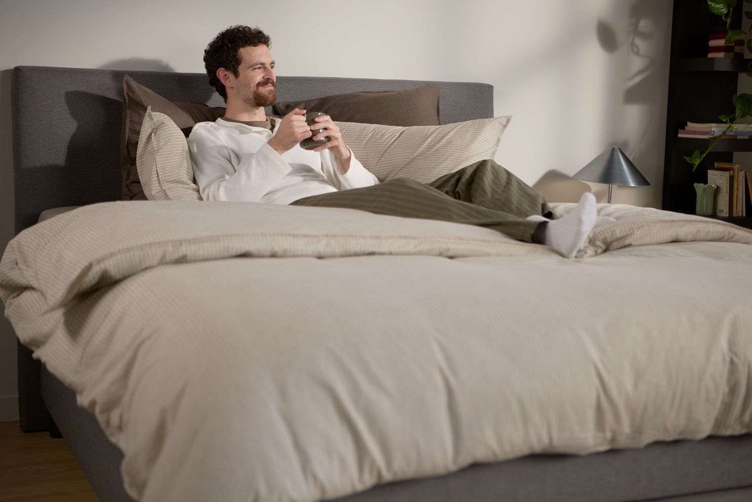 Iemand zit op een bed met een zacht, gestoffeerd hoofdbord, meerdere laagjes kussens en een dekbed in neutrale tinten, in een zacht verlichte slaapkamer.