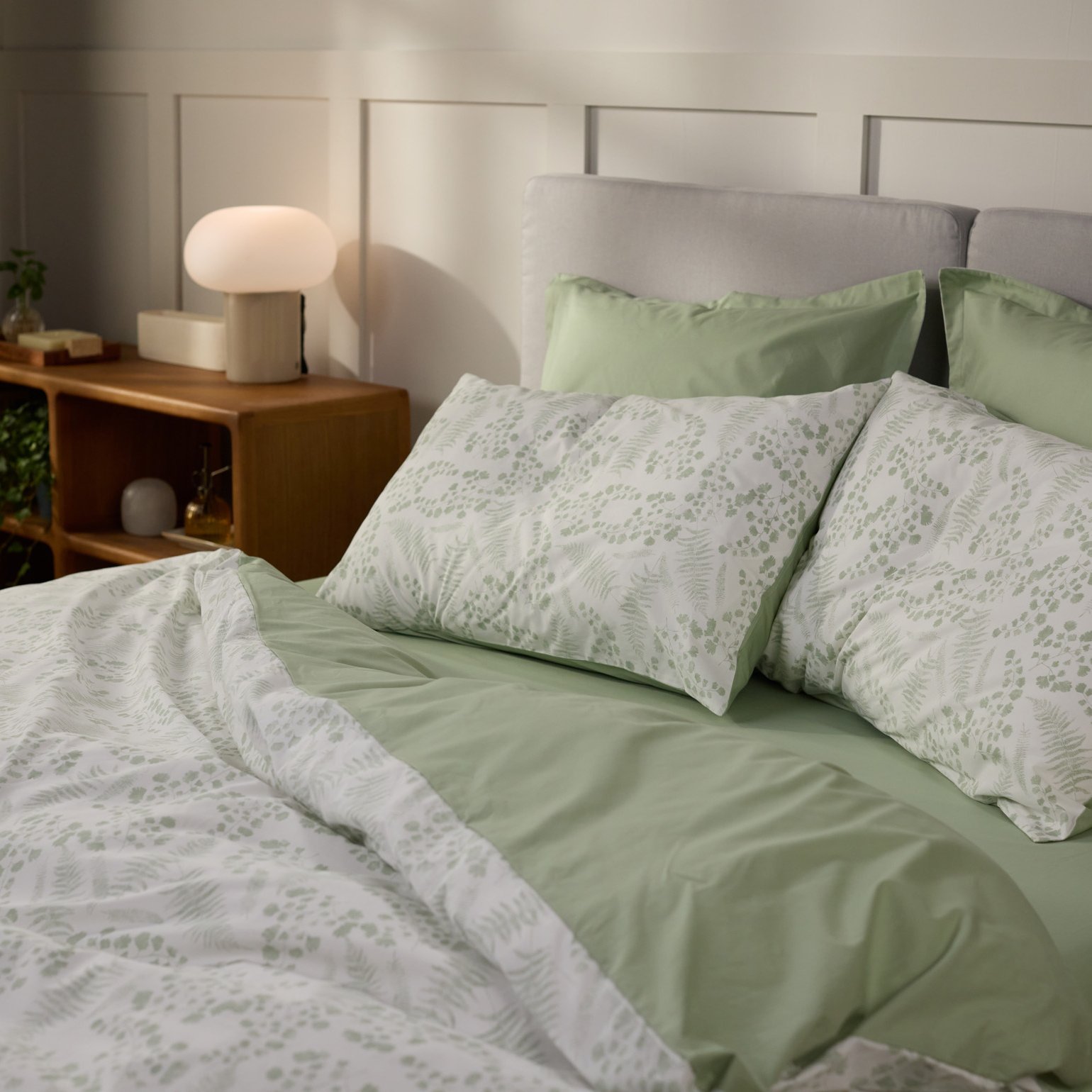 Opgemaakt bed met lichtgroen beddengoed en kussens met een botanisch patroon, naast een houten nachtkastje met lamp en plant in een lichte slaapkamer.