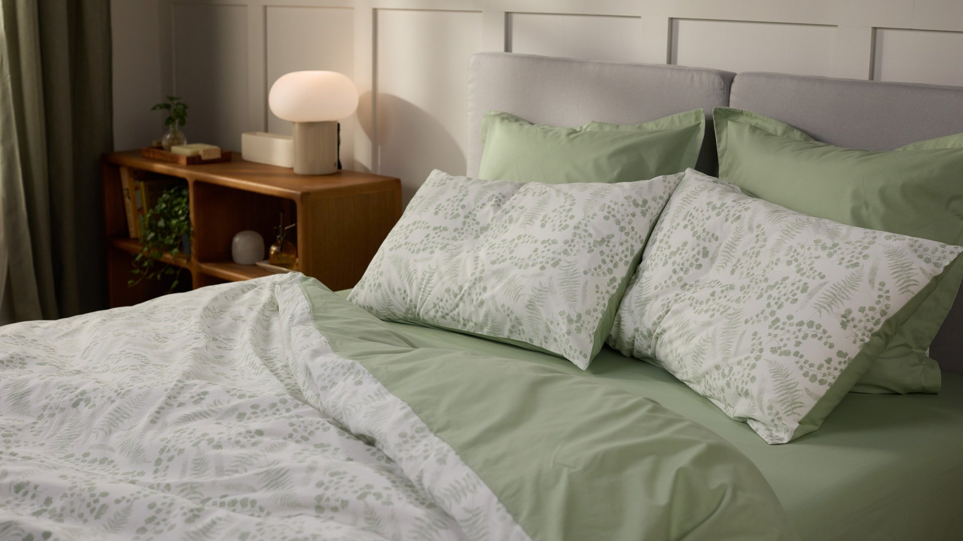 Opgemaakt bed met lichtgroen beddengoed en kussens met een botanisch patroon, naast een houten nachtkastje met lamp en plant in een lichte slaapkamer.