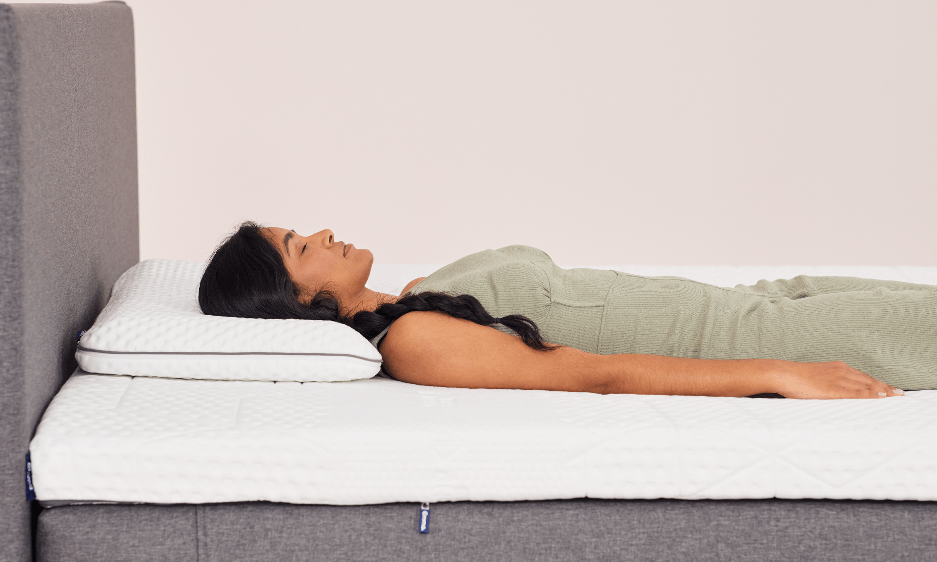 Personne allongée sur le dos sur un lit avec surmatelas blanc texturé et oreiller assorti, cadre de lit en tissu gris, dans une chambre lumineuse. La personne porte une robe verte côtelée.